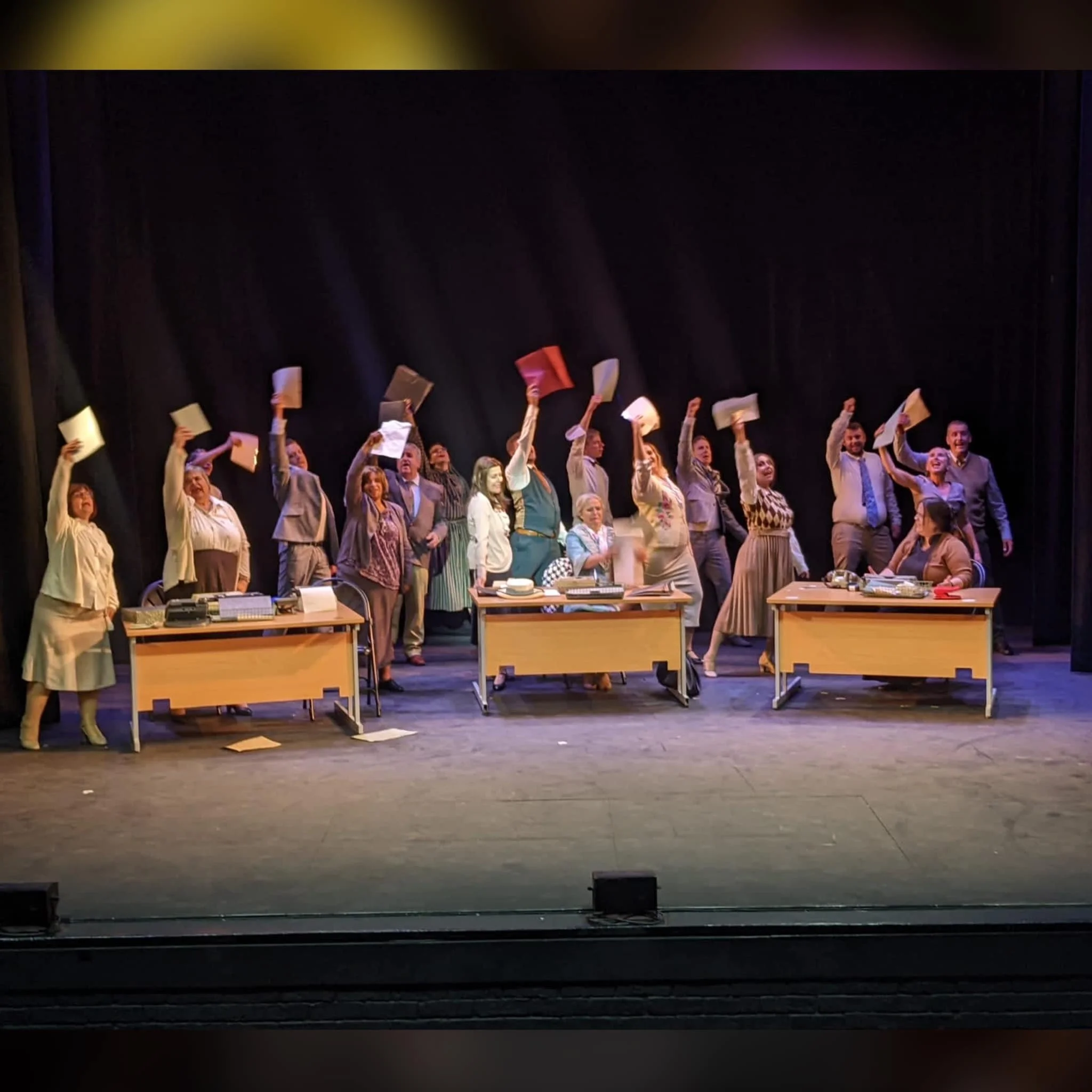 Group of actors on stage at the end of a play, waving papers or scripts in the air, with desks and props on stage.