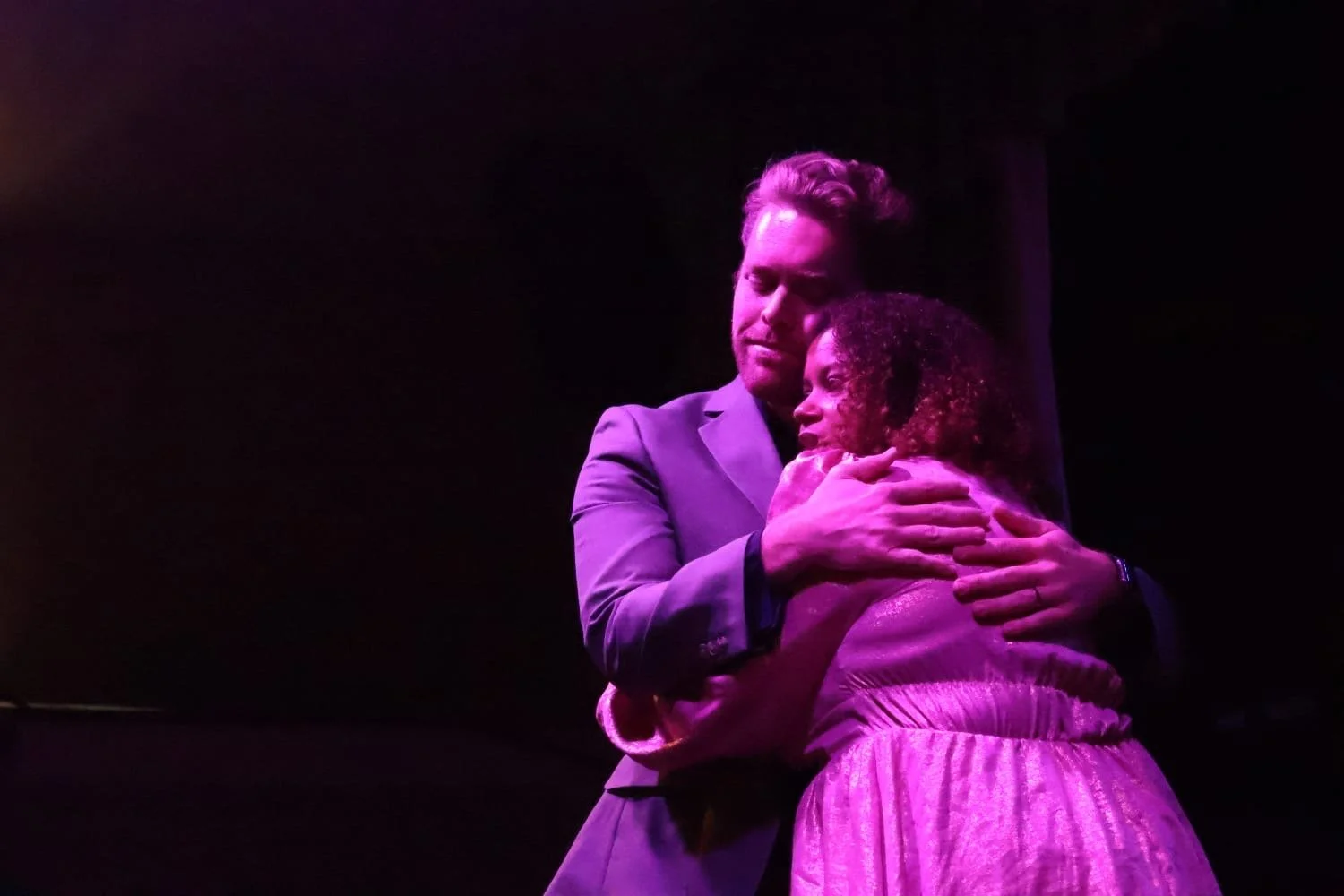 A man and woman embracing on stage with purple lighting, the man has blond hair and a suit, the woman has curly hair and a striped dress.