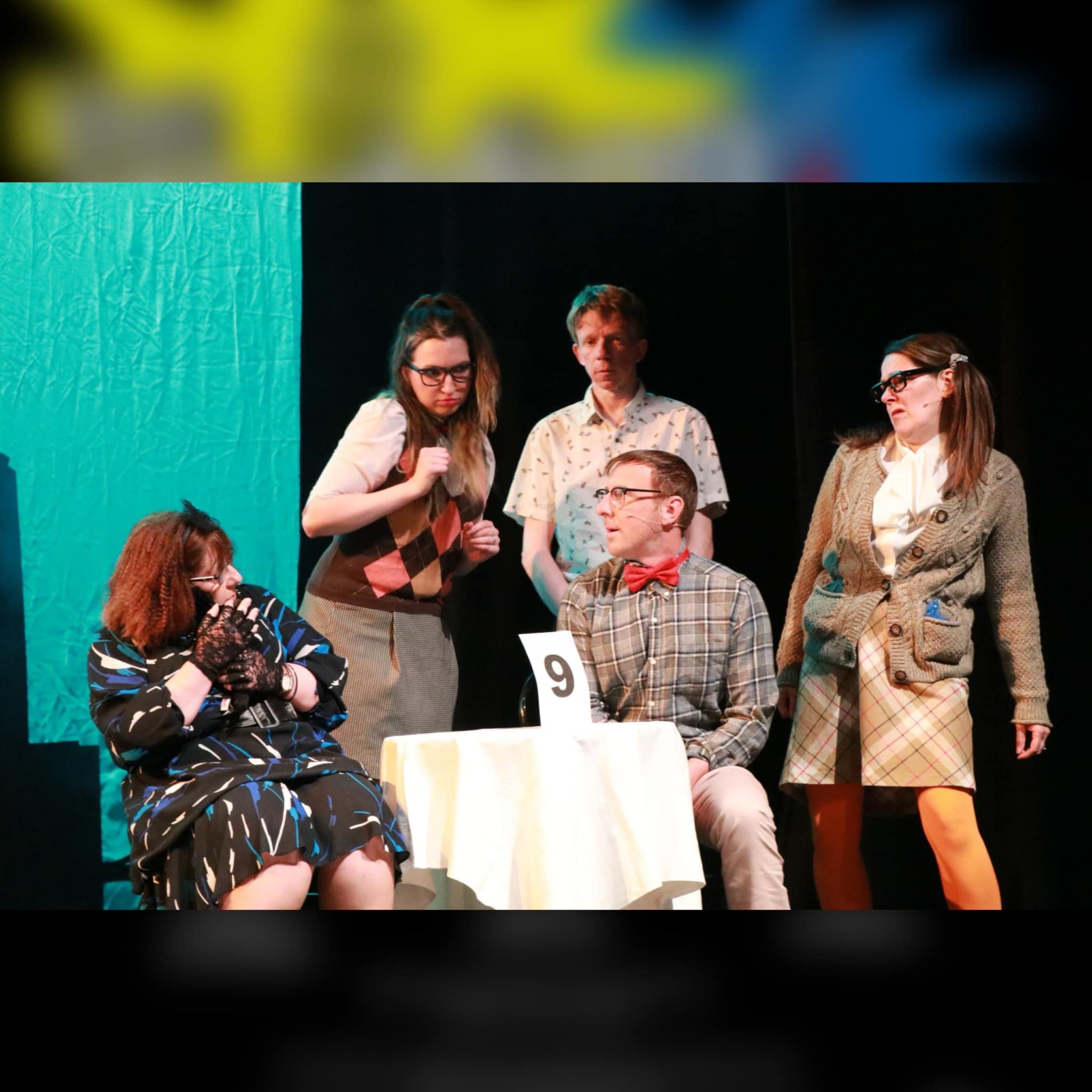 Photograph of five actors on stage, five portraying characters in a scene, with a table marked with the number 9 in the center. The scene appears dramatic, with various facial expressions and costumes.