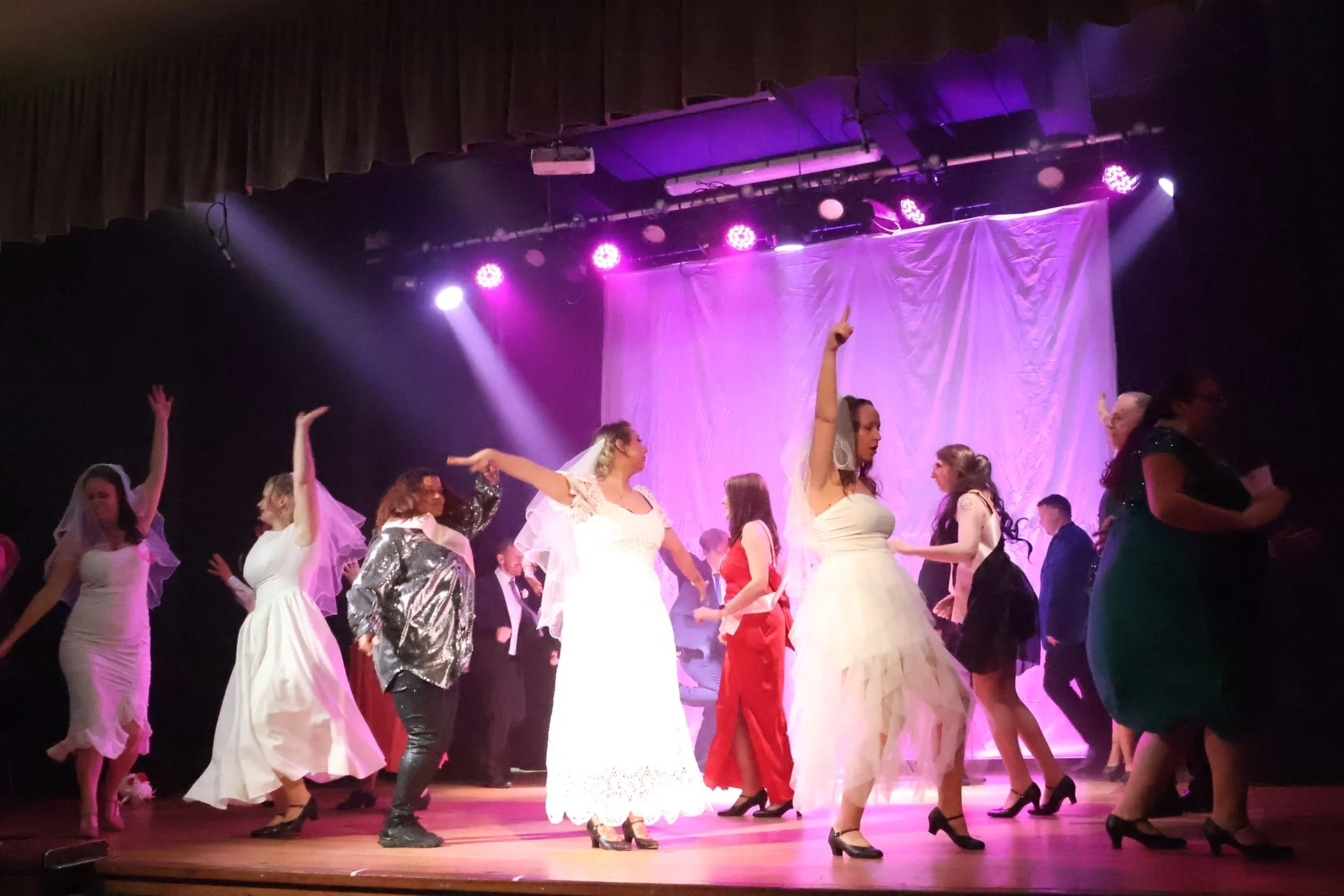 Group of women dancing on stage during a performance, some wearing white wedding dresses, in front of a purple-lit curtain.