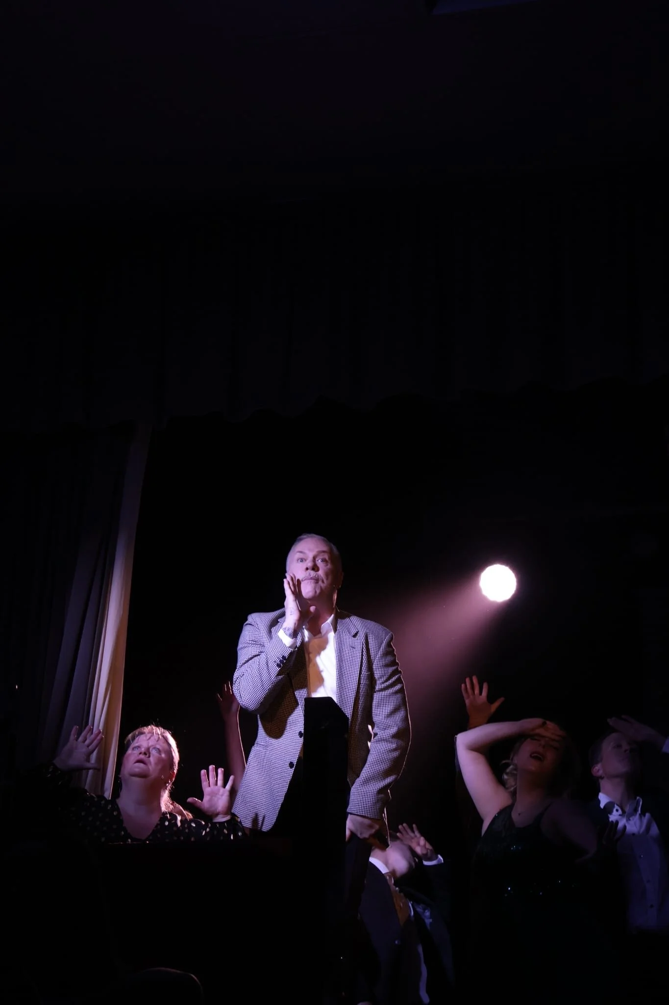 A man in a checkered blazer standing on stage with other performers and a spotlight shining behind him.