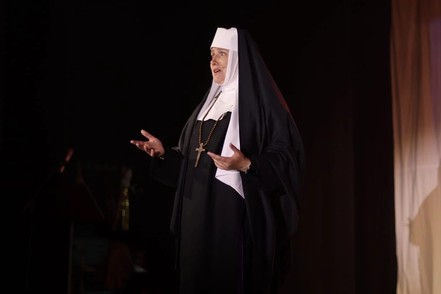 A woman dressed as a nun is speaking or performing on a stage, wearing a black and white habit and a large cross necklace, with a dark background and stage lighting.