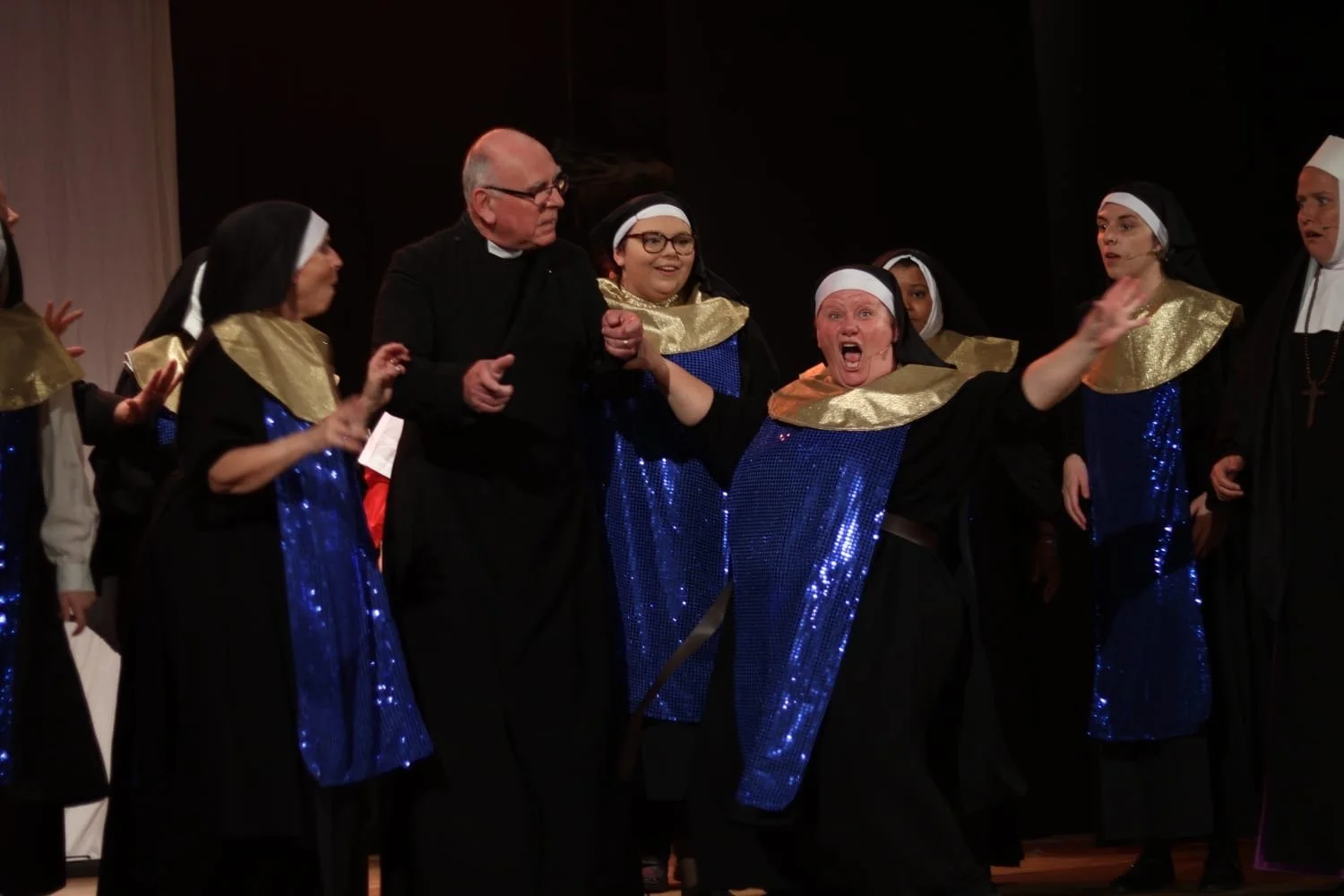 Group of performers dressed as nuns in costumes with blue and gold sequined robes, on stage during a theatrical performance.
