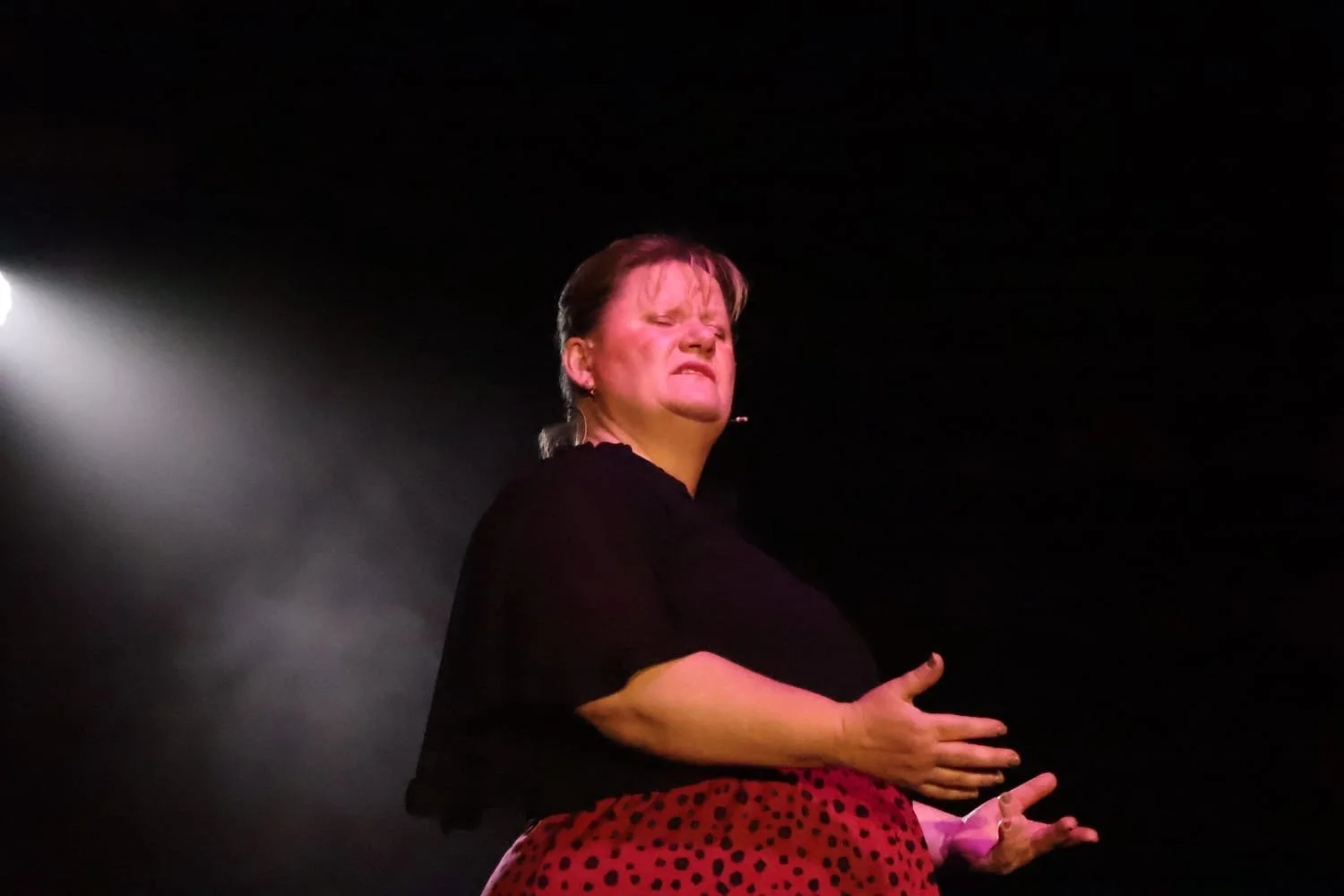 A woman performing on stage, squinting with a facial expression of effort or intensity, against a dark background with a spotlight shining from the left.
