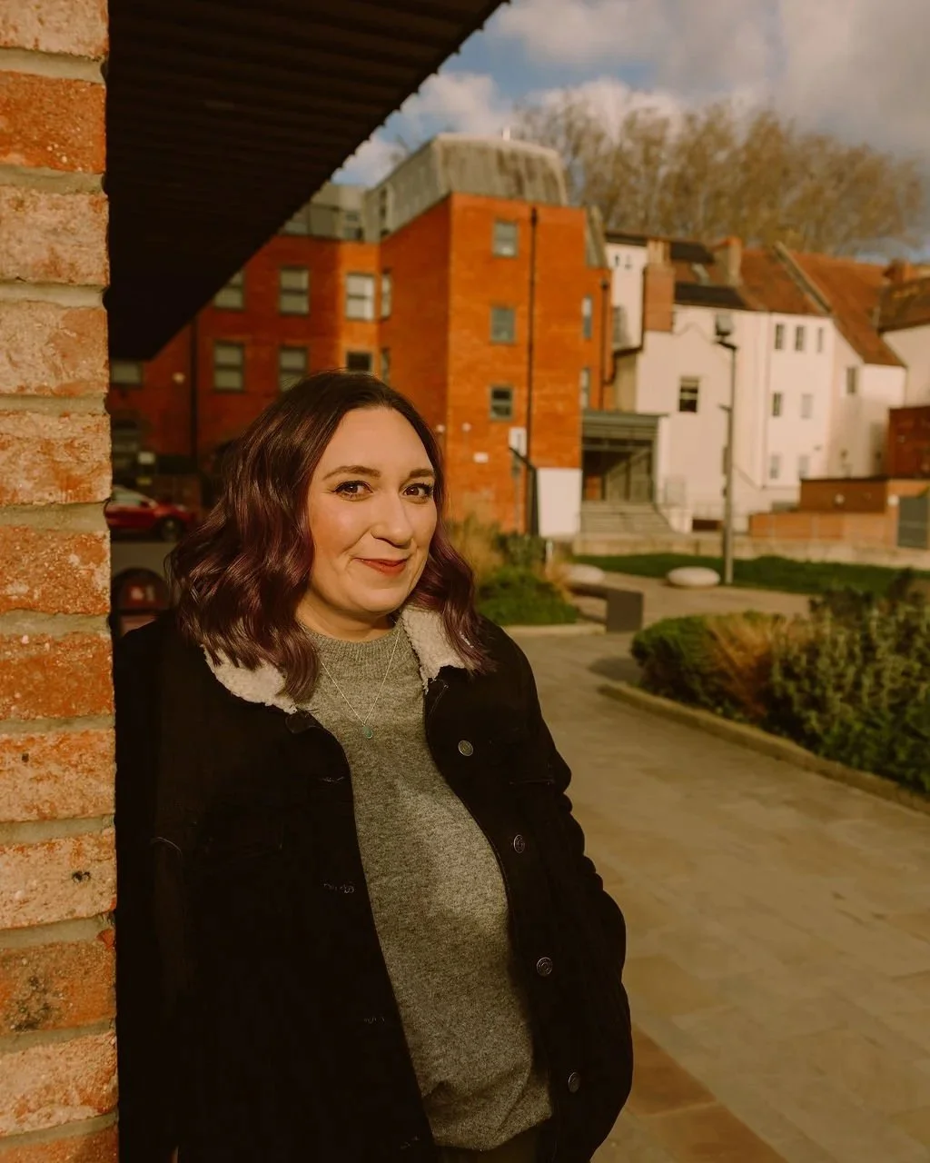 A woman with shoulder-length wavy brown hair standing outdoors near a brick wall, smiling at the camera with residential buildings and a pathway in the background during what appears to be late afternoon or early evening.