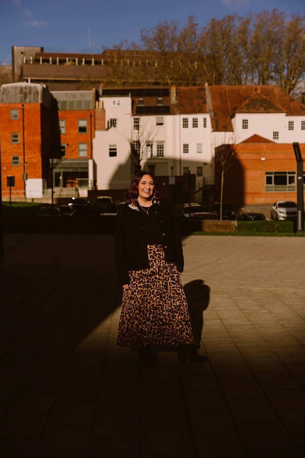 A woman standing on a paved area in front of buildings with a mix of white and red brick facades, smiling. The scene is brightly lit with sunlight casting shadows.