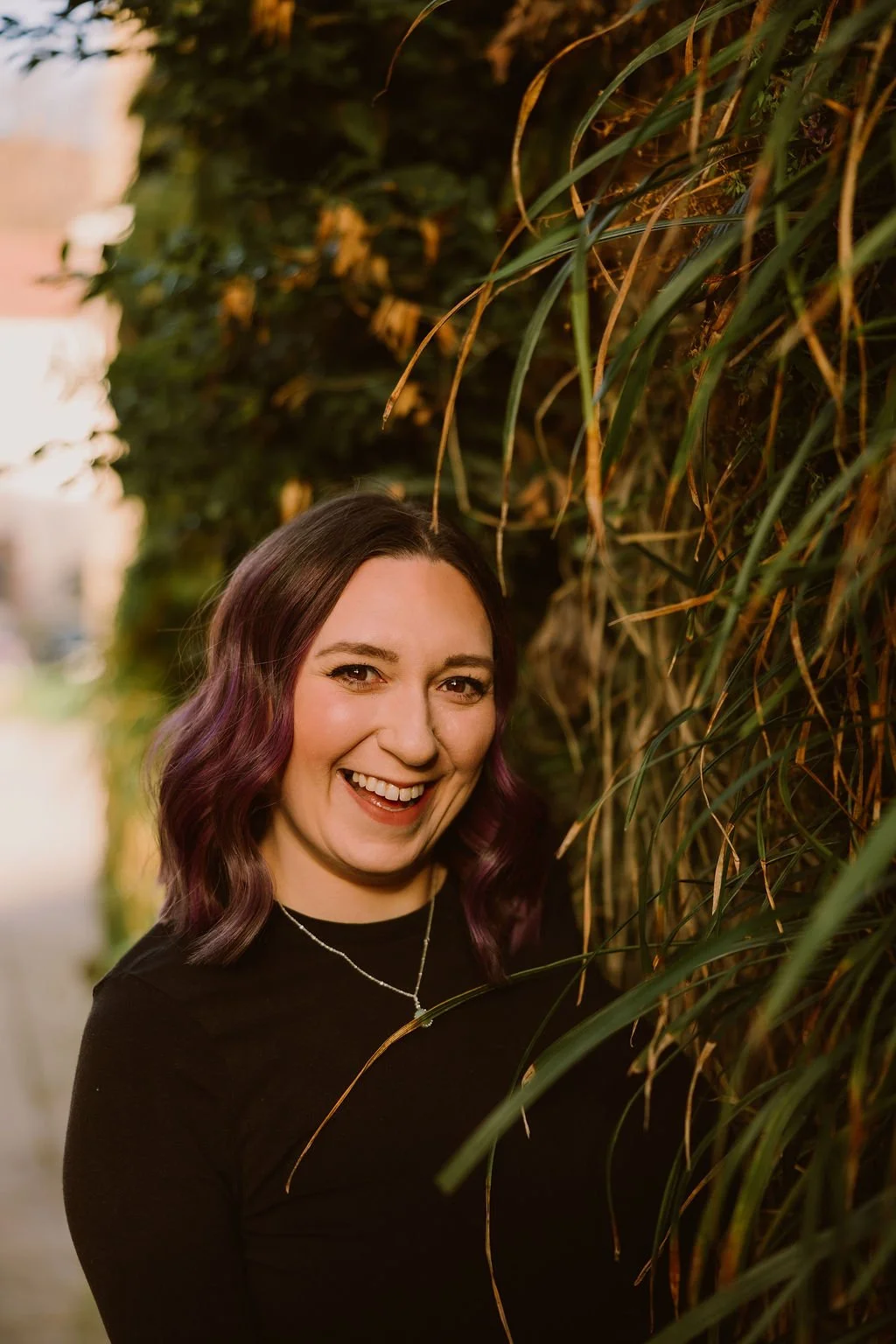 A woman smiling and posing outdoors next to tall grass or plants.