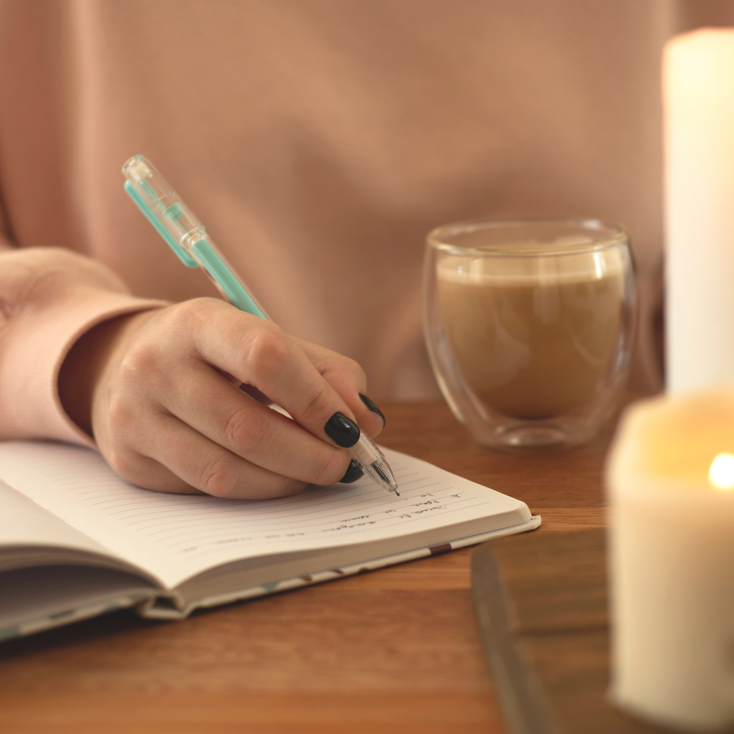A person writing in a notebook with a pen while sitting at a wooden table, with a glass of coffee and a lit candle nearby.