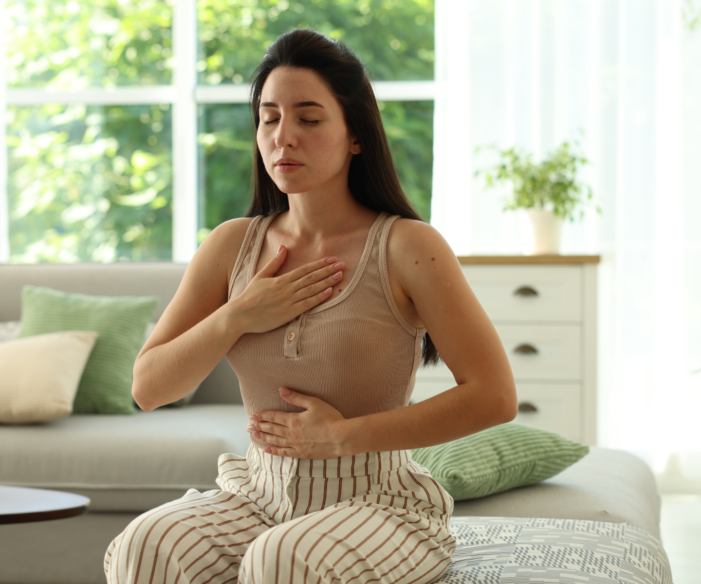 A woman sitting cross-legged on a bed, experiencing chest discomfort, holding her chest and stomach, with eyes closed in a bright bedroom.