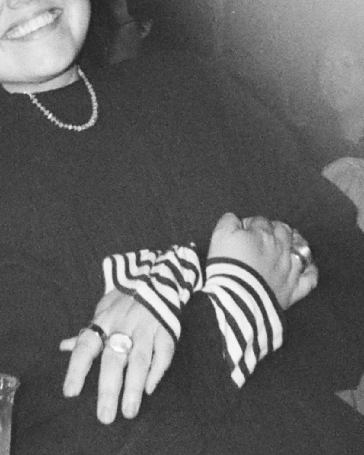 Close-up black and white photo of a person's hand with rings, wearing striped long sleeves, resting on a dark surface, partially visible face with a pearl necklace, and a smile, in the background.
