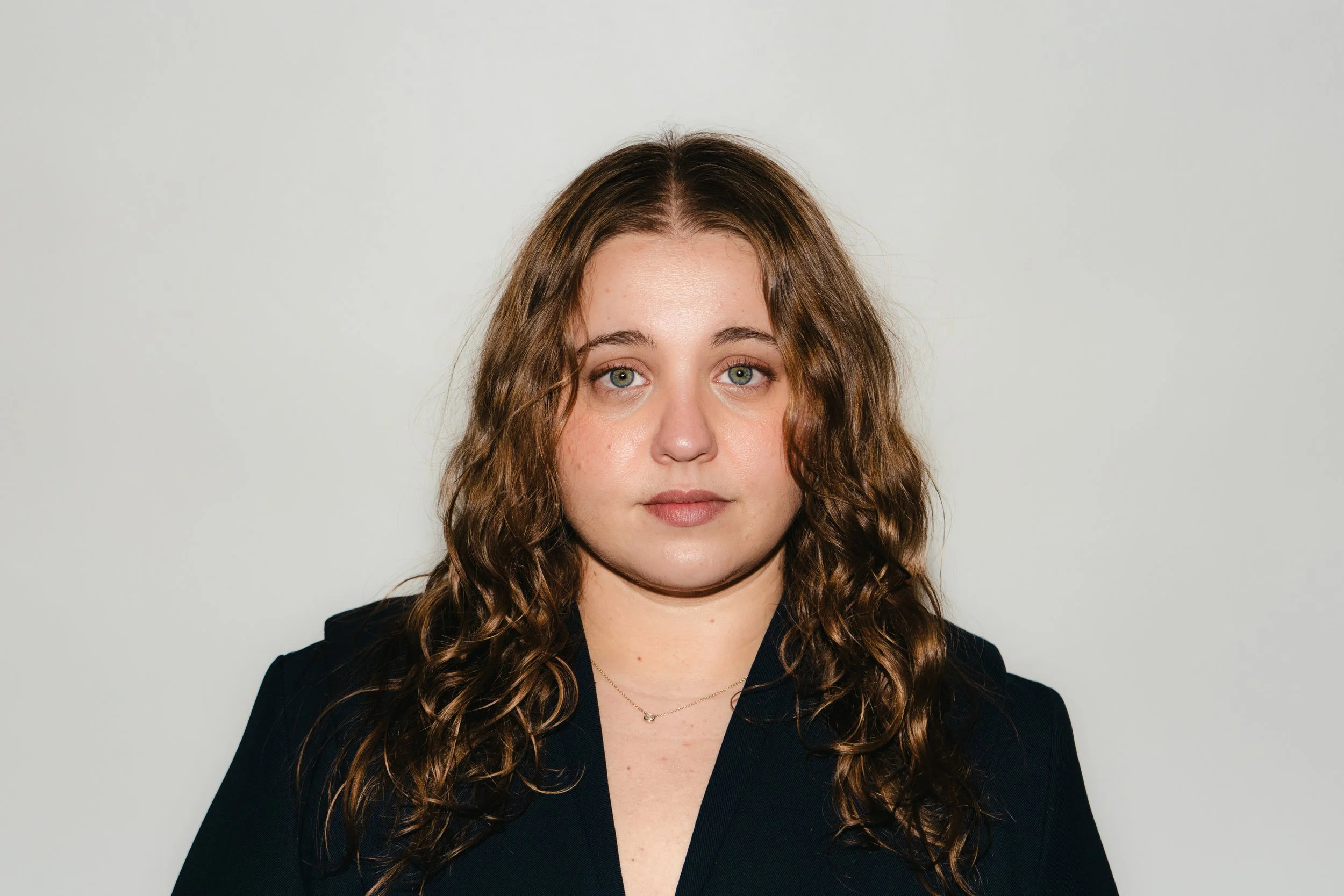 A woman with wavy brown hair wearing a black top and a delicate necklace poses against a plain light gray background.