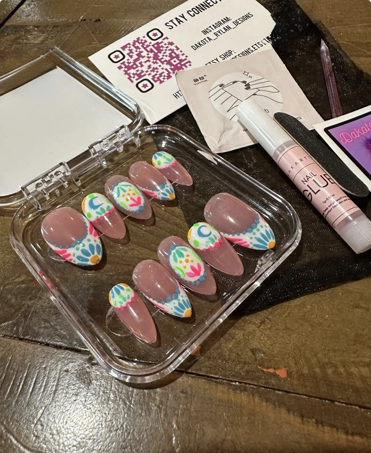 A set of decorated artificial nails with colorful, floral and abstract patterns, placed in a clear plastic box on a wooden table, along with nail art supplies and printed instructions.