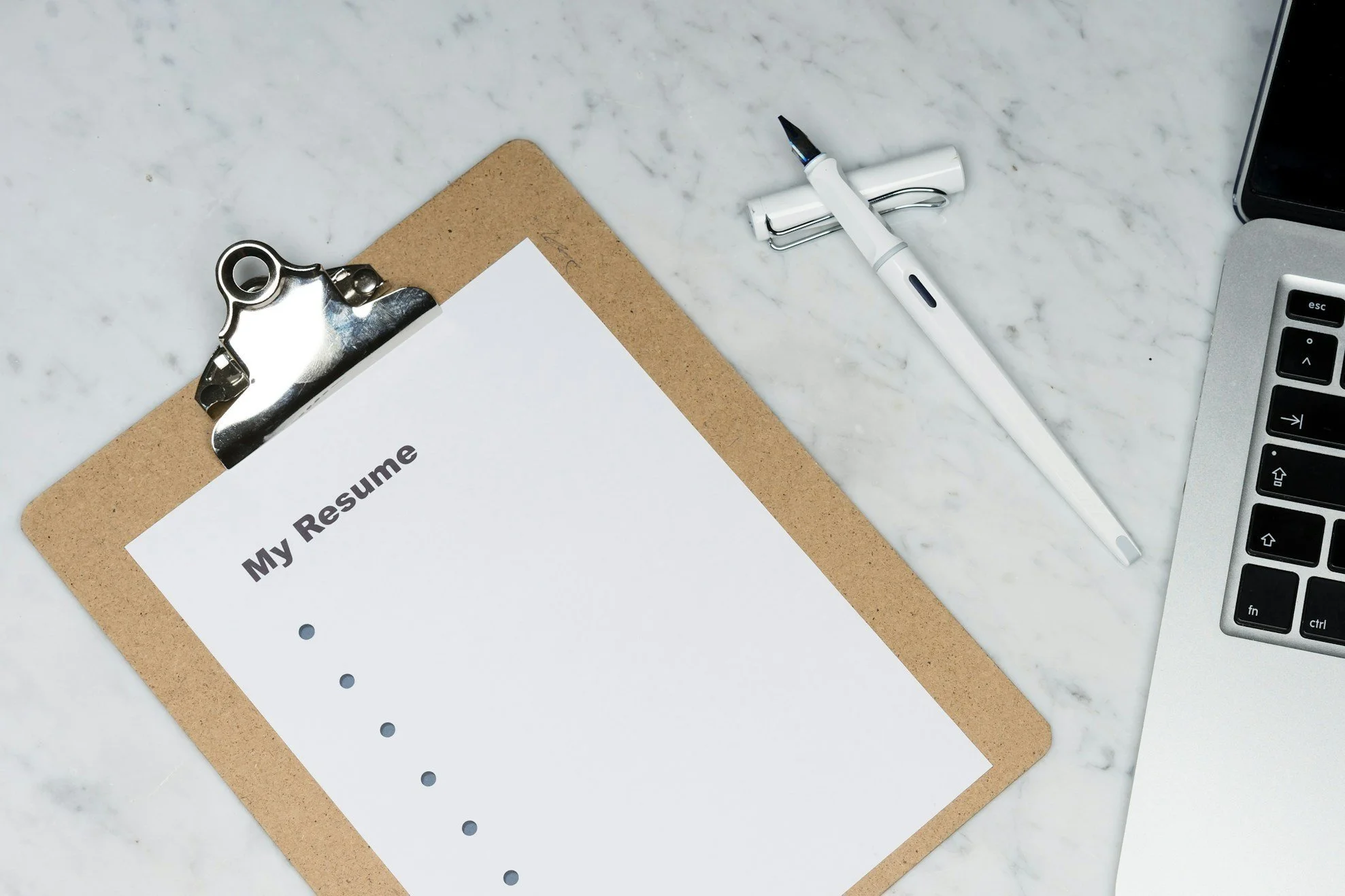 A resume on a clipboard with a pen, a laptop, and a smartphone on a marble surface.