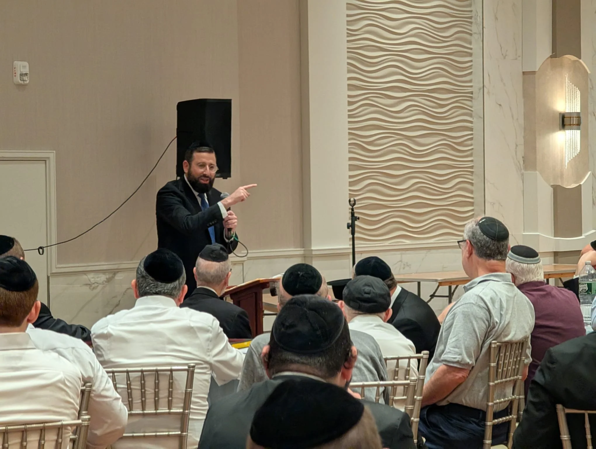 Rabbi Daniel Glatstein, holding a microphone and pointing, is speaking at an event with an audience of men, many wearing kippahs, seated in rows of chairs in a large, well-lit room with decorative wall panels and a speaker system.