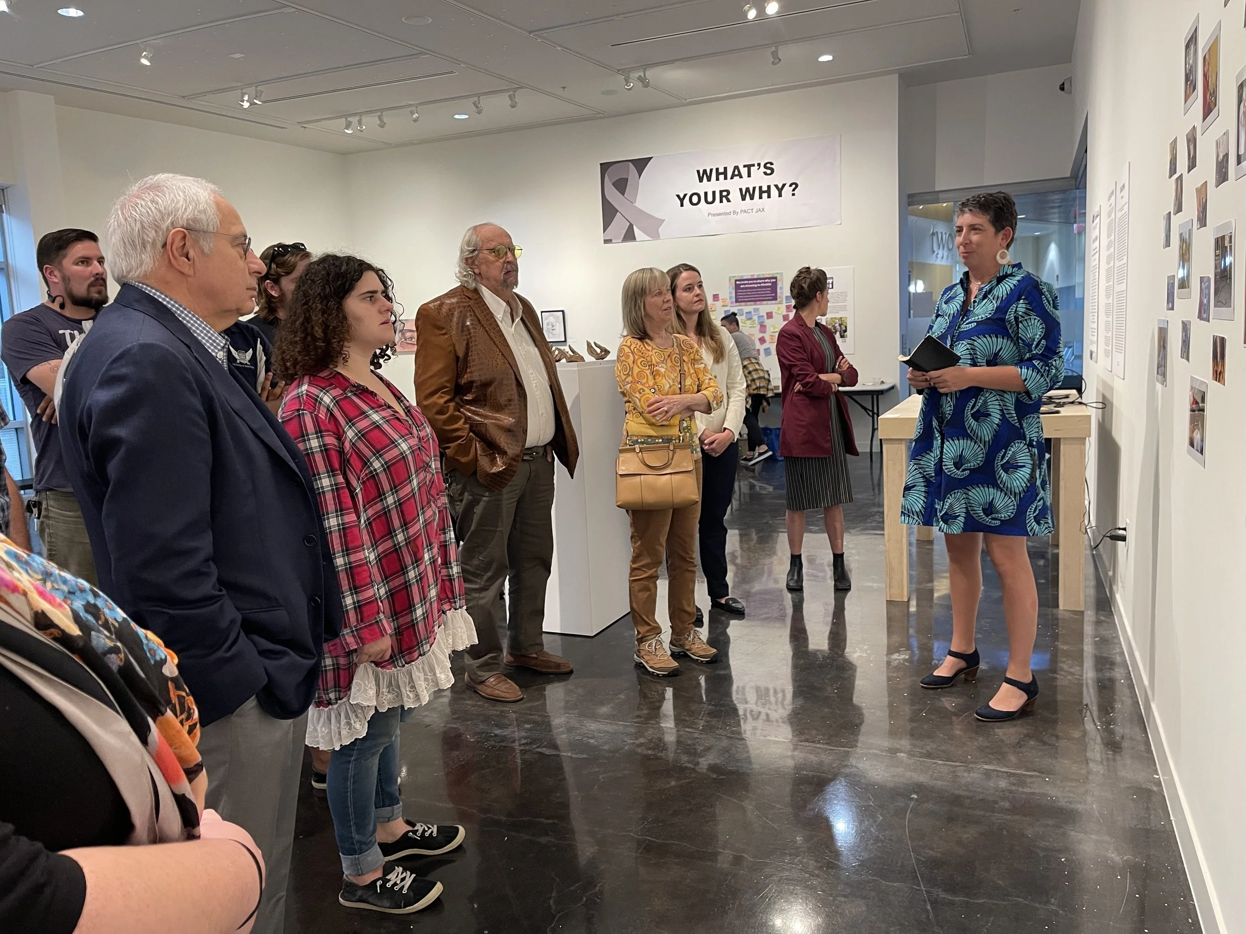 A woman in a blue patterned dress is giving a talk to a group of people at an art gallery. The audience is standing and attentively listening. There is a large sign on the wall behind her that says, "WHAT'S YOUR WHY?"
