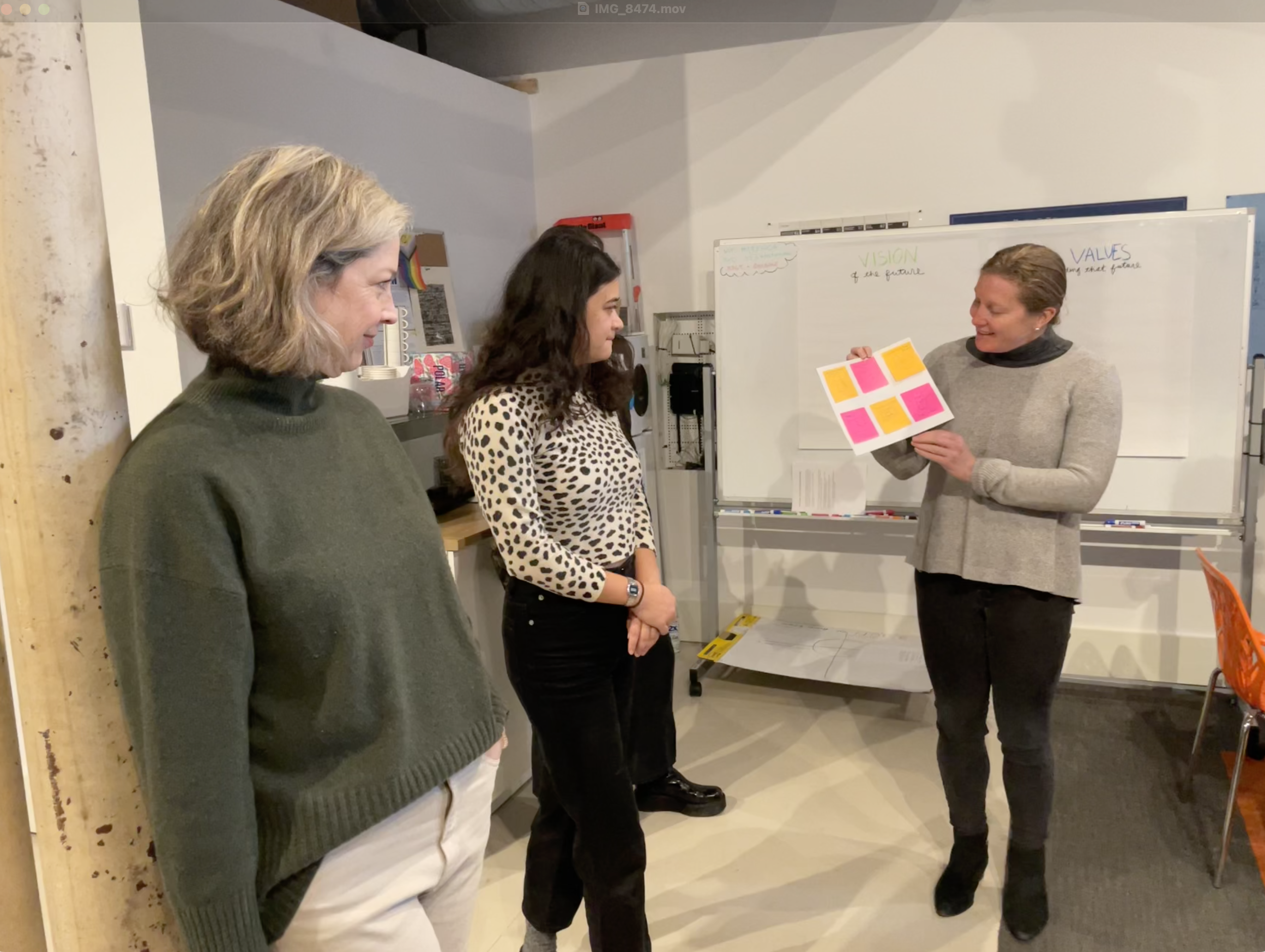 A woman presenting sticky notes on a whiteboard to two women in a meeting room.
