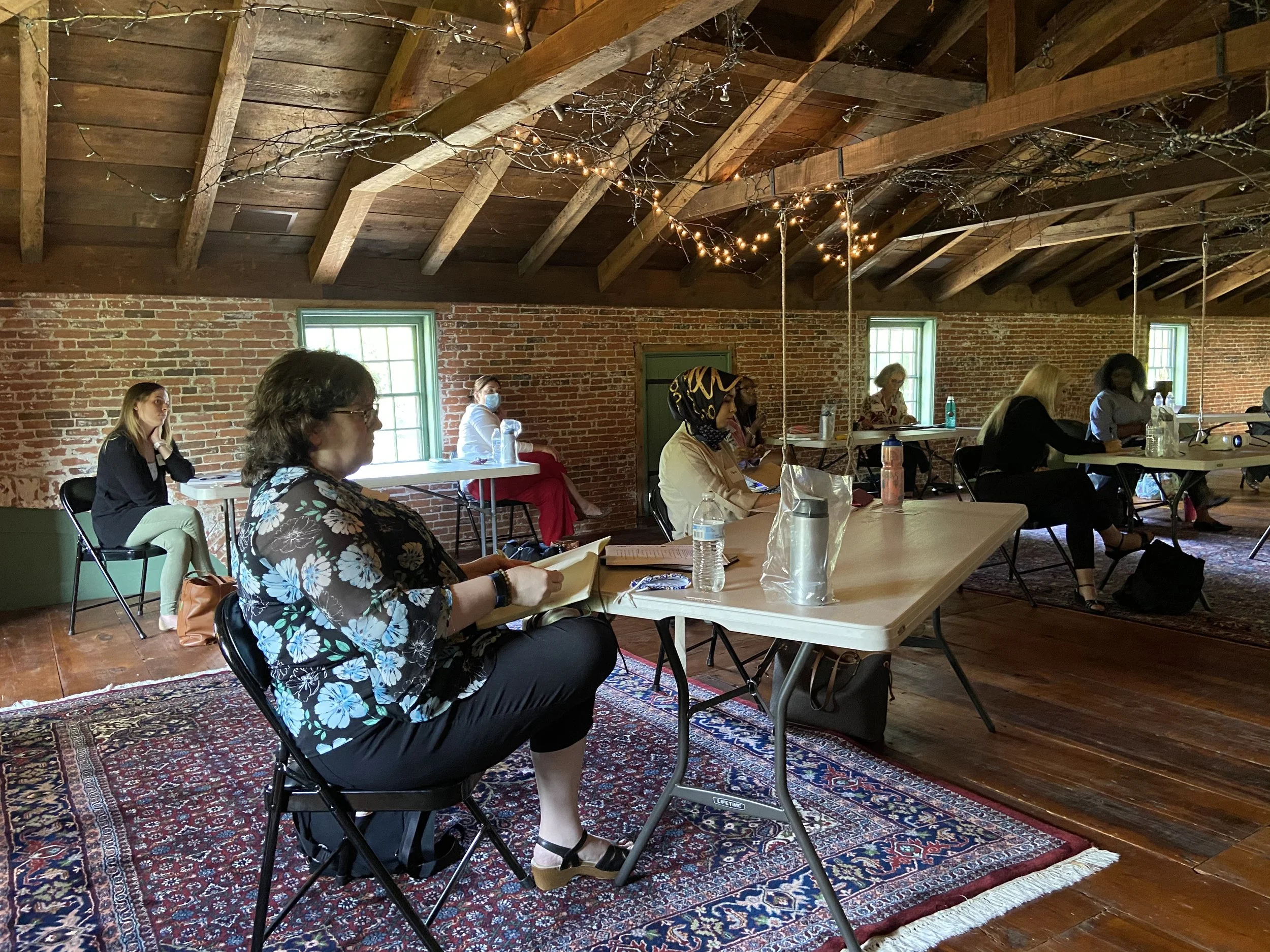 Group of women sitting in socially distanced positions in a rustic brick-walled room with a wooden ceiling, fluorescent lighting, and windows, participating in a workshop or meeting. Most are wearing masks and have papers, bottles, and water on the tables.