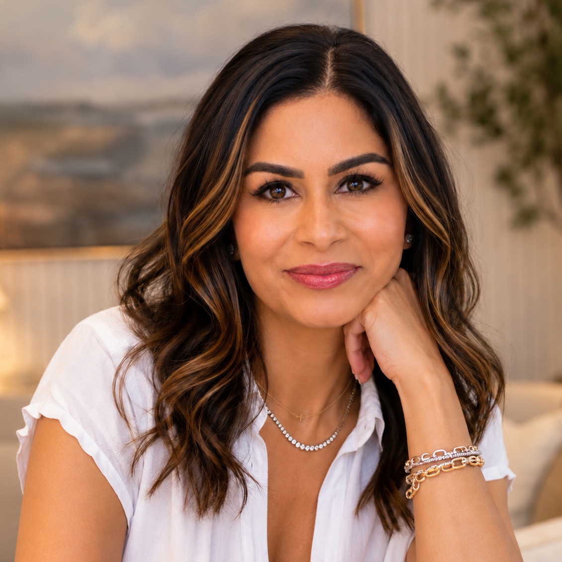 Portrait of a smiling woman with long wavy brown hair, wearing a white blouse, layered necklaces, and bracelets.