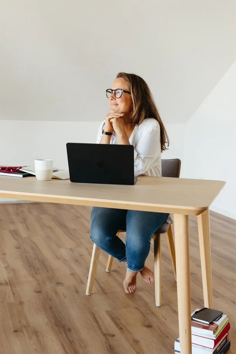 Eine Frau in legerer Kleidung sitzt mit ihrem Laptop an einem Holztisch und schaut verträumt zur Seite.