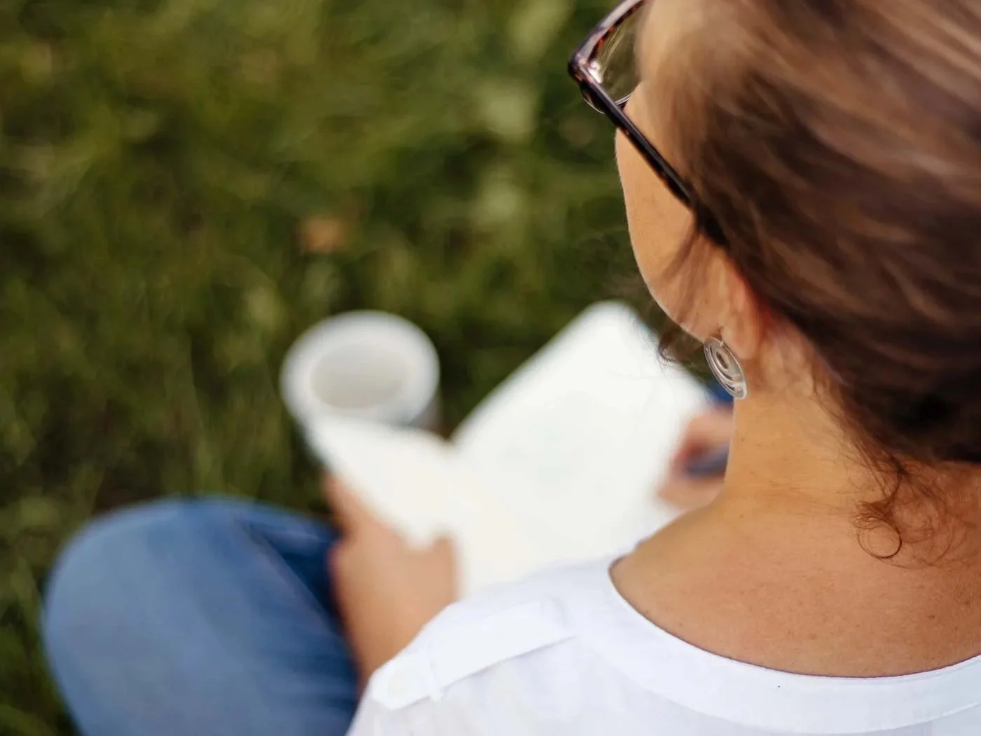 Eine Frau mit Brille und einem Notizbuch in der Hand sitzt im Gras und schaut weg von der Kamera.