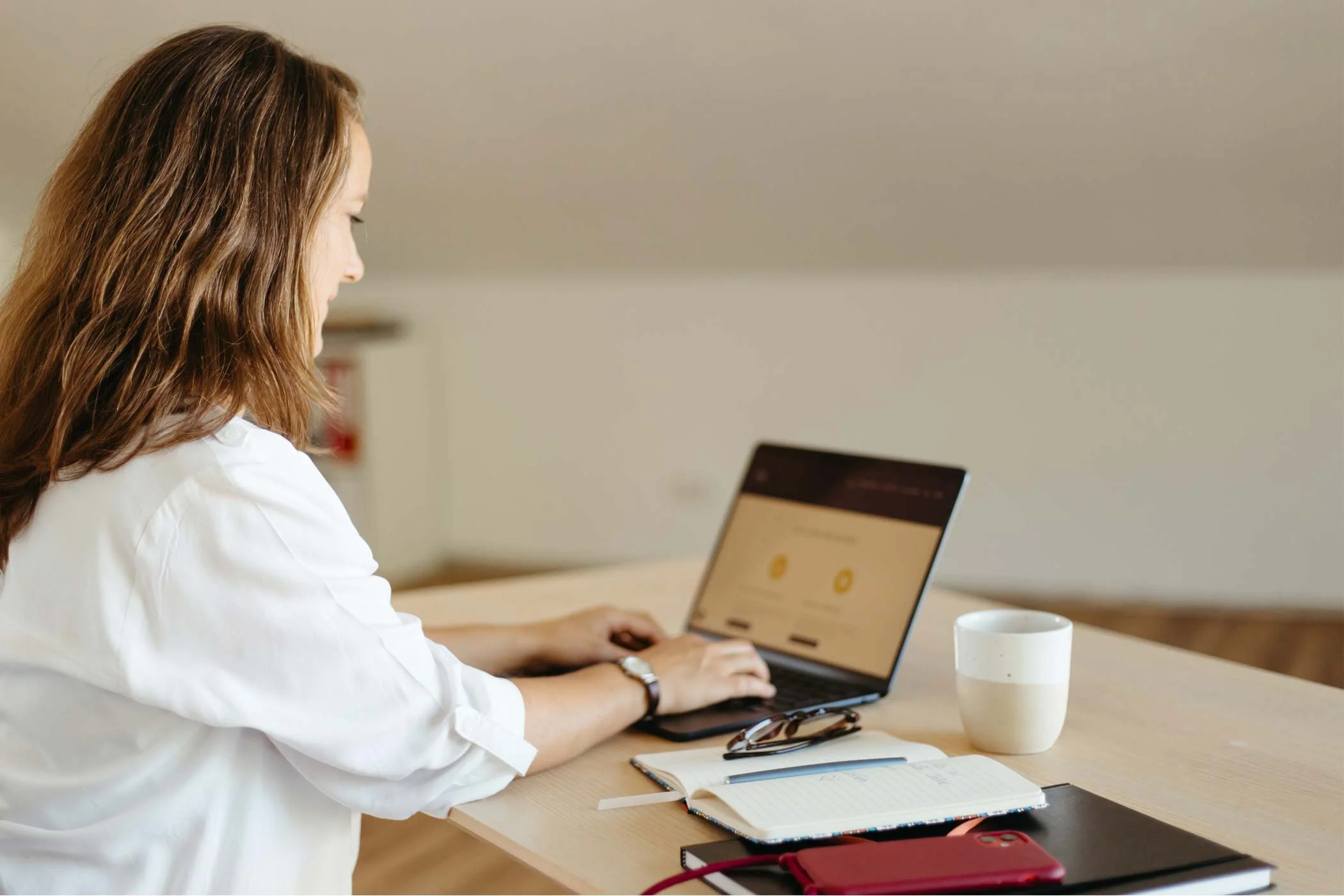 Eine Frau in weißer Bluse sitzt am Tisch und arbeitet an einem Laptop.