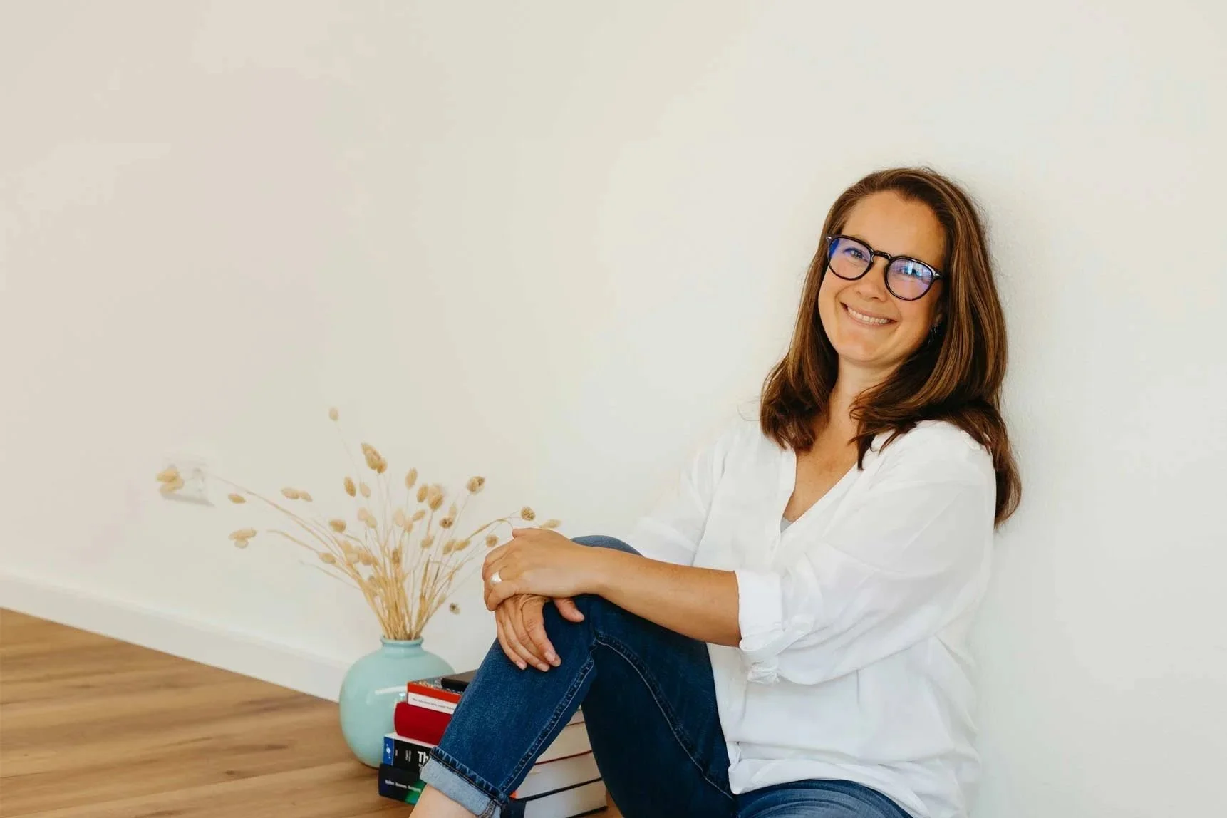 Eine lächelnde Frau mit Brille sitzt auf dem Boden in einem hellen Raum, neben einer Vase mit Trockenblumen und einem Stapel Bücher.