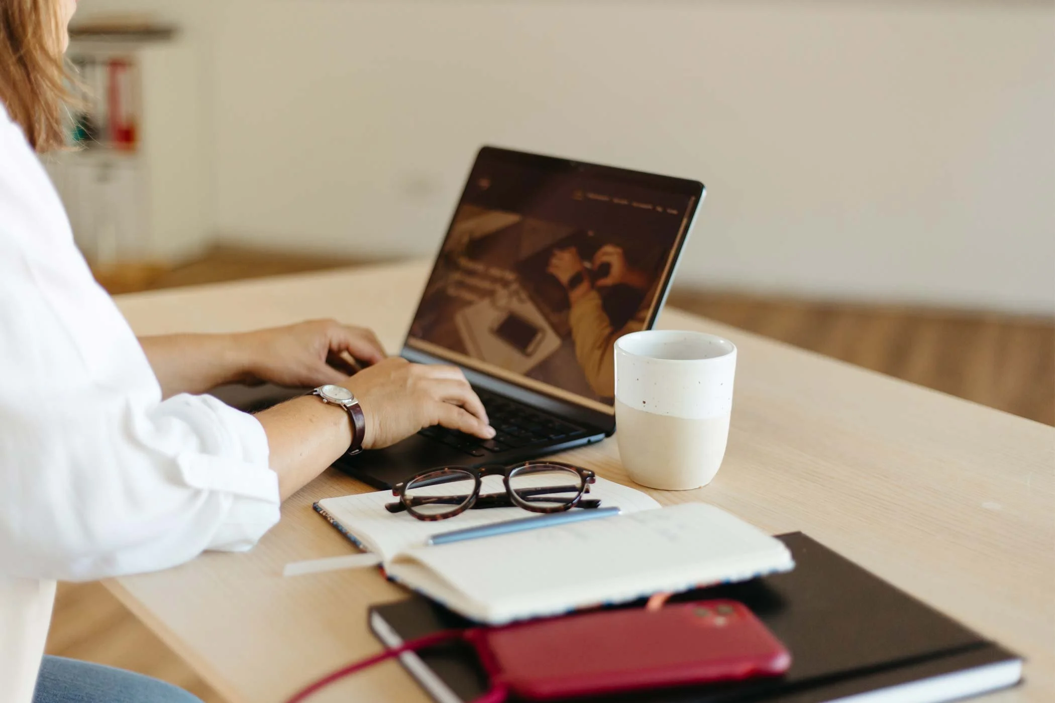 Eine Frau in weißer Bluse arbeitet an ihrer Website. Daneben liegen ein Notizbuch, ein Handy und eine Brille.