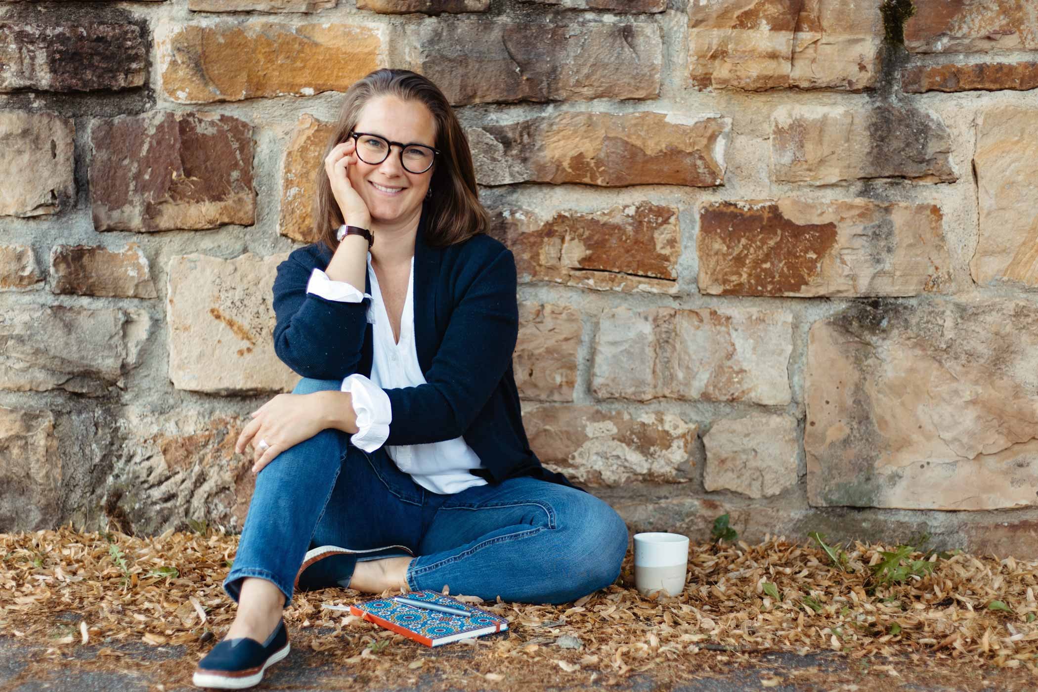 Eine Frau mit Brille sitzt vor einer Mauer und lächelt in die Kamera.
