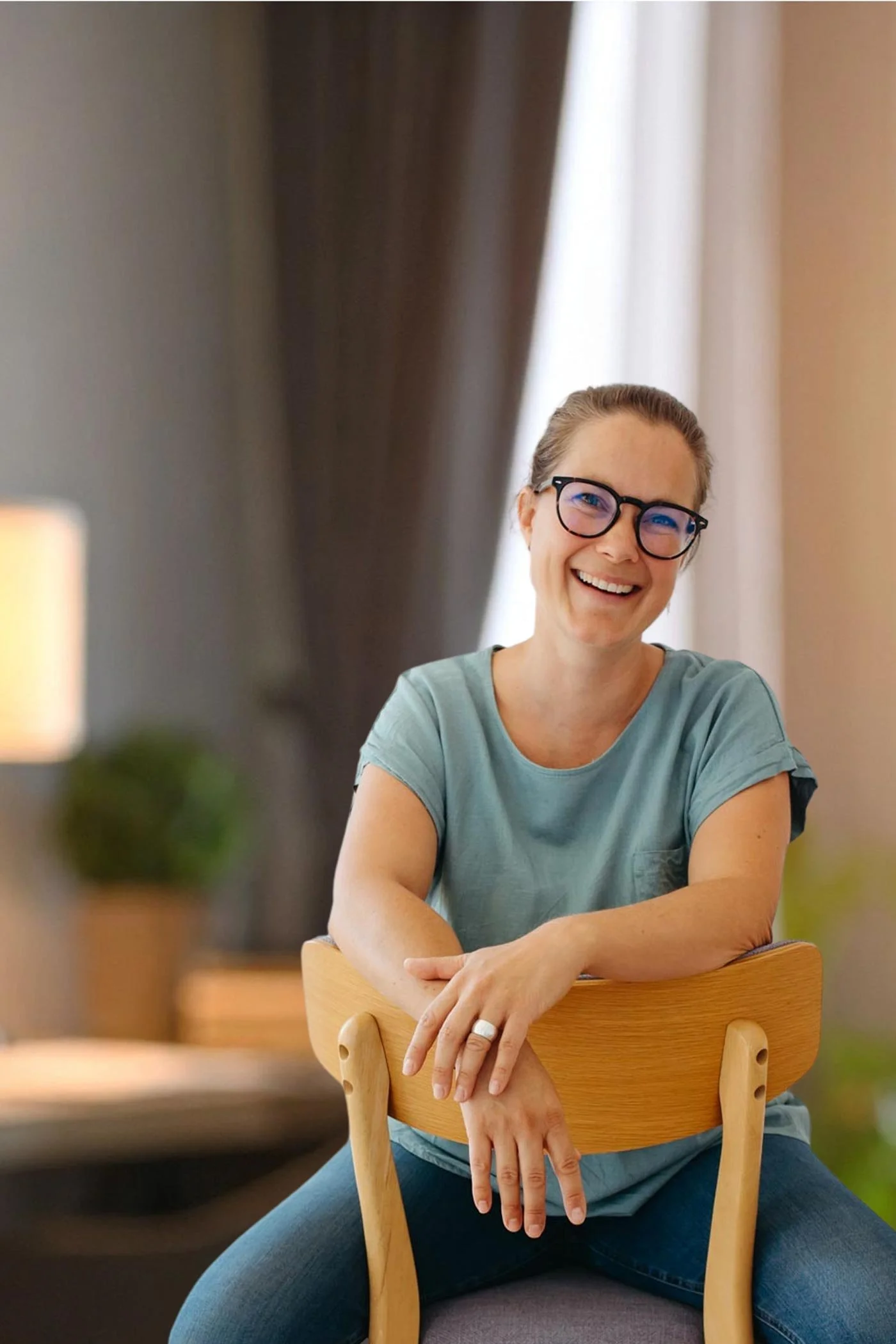 Eine Frau mit einer grünen kurzen Bluse, Jeans und Brille sitzt auf einem Holzstuhl und lacht in die Kamera.