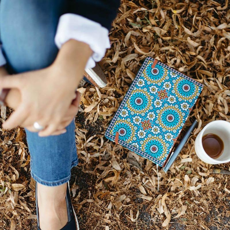 Eine Frau in Jeans sitzt auf dem Boden und neben ihr liegt ein buntes Notizbuch.