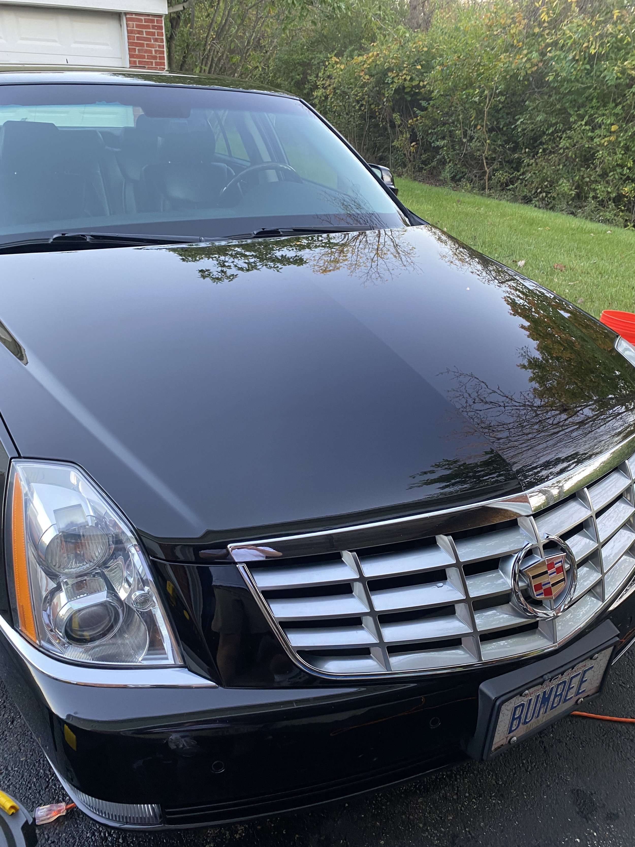 A black Cadillac car parked outdoors in a driveway with a grass lawn and trees in the background. The car has a custom license plate that reads 'BUMBE'.