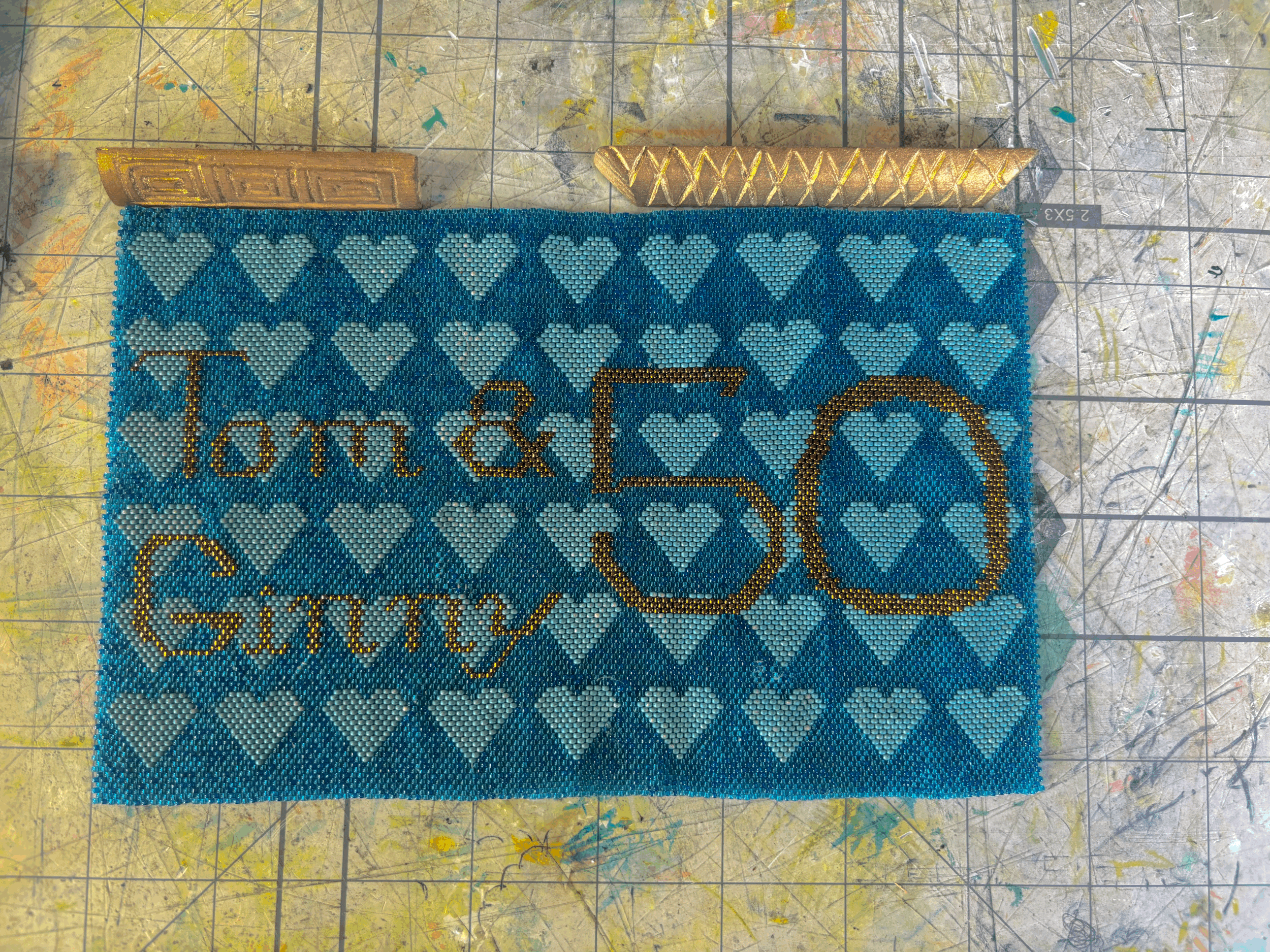 A blue tapestry with a light blue heart pattern. The tapestry says “Tom & Ginny 50” in gold lettering to commemorate a 50th wedding anniversary.