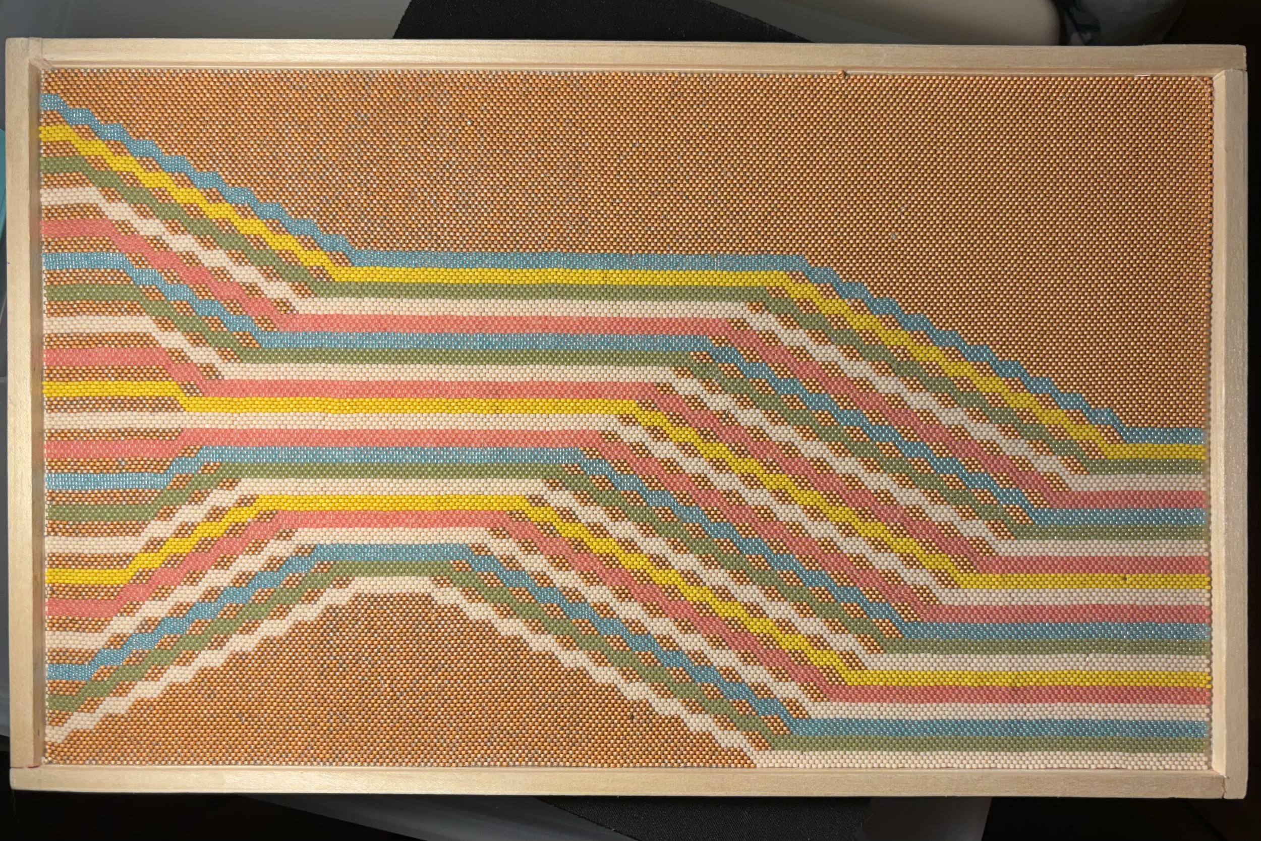 An abstract geometric beaded tapestry with ribbons of blue, yellow, green, white, and pink cascading in formation, like staircases, from left to right, against a gold background.
