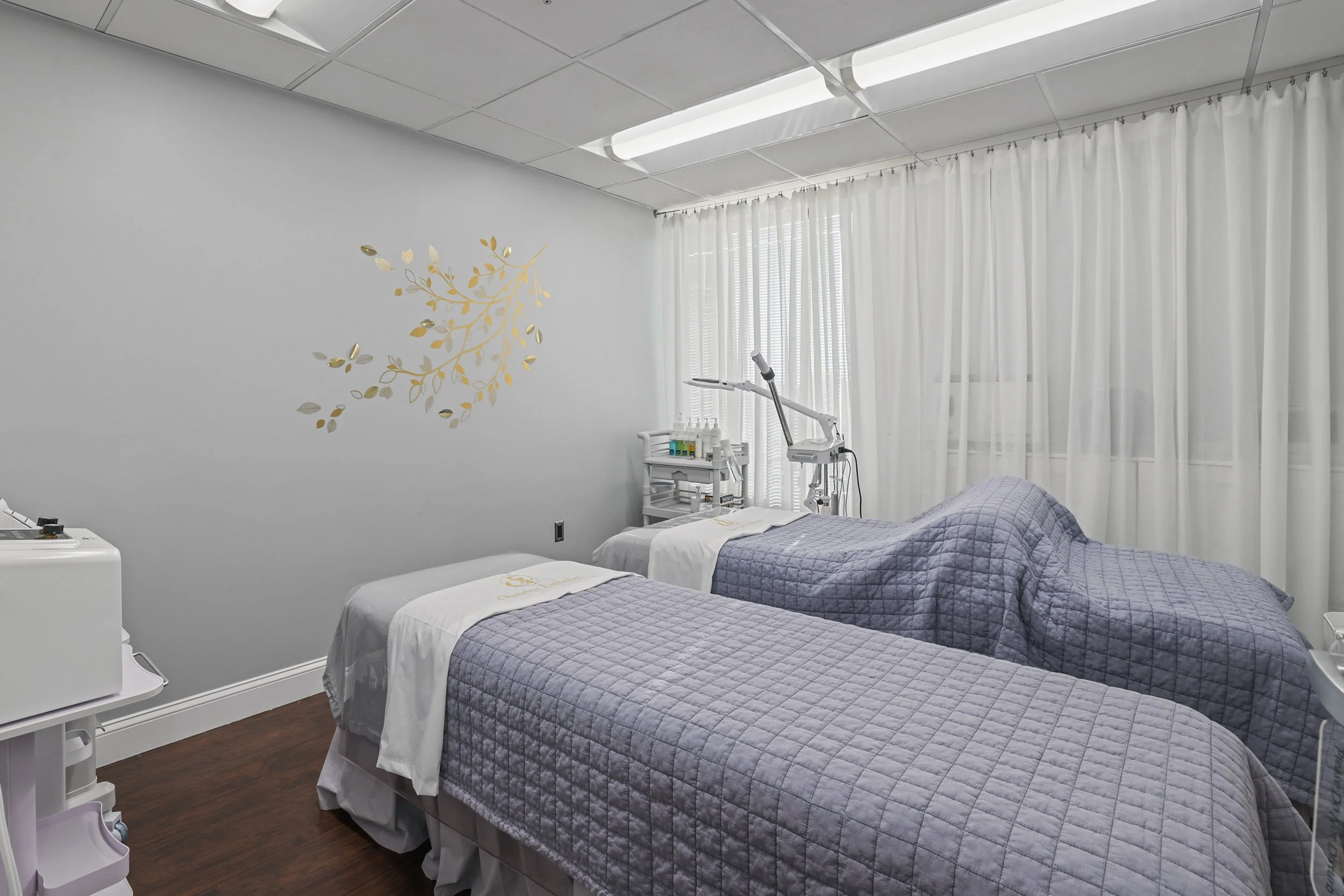 Treatment room with two beds, each covered with a gray quilt. The room is bright, with white curtains and a wall decorated with gold and silver leaf design.