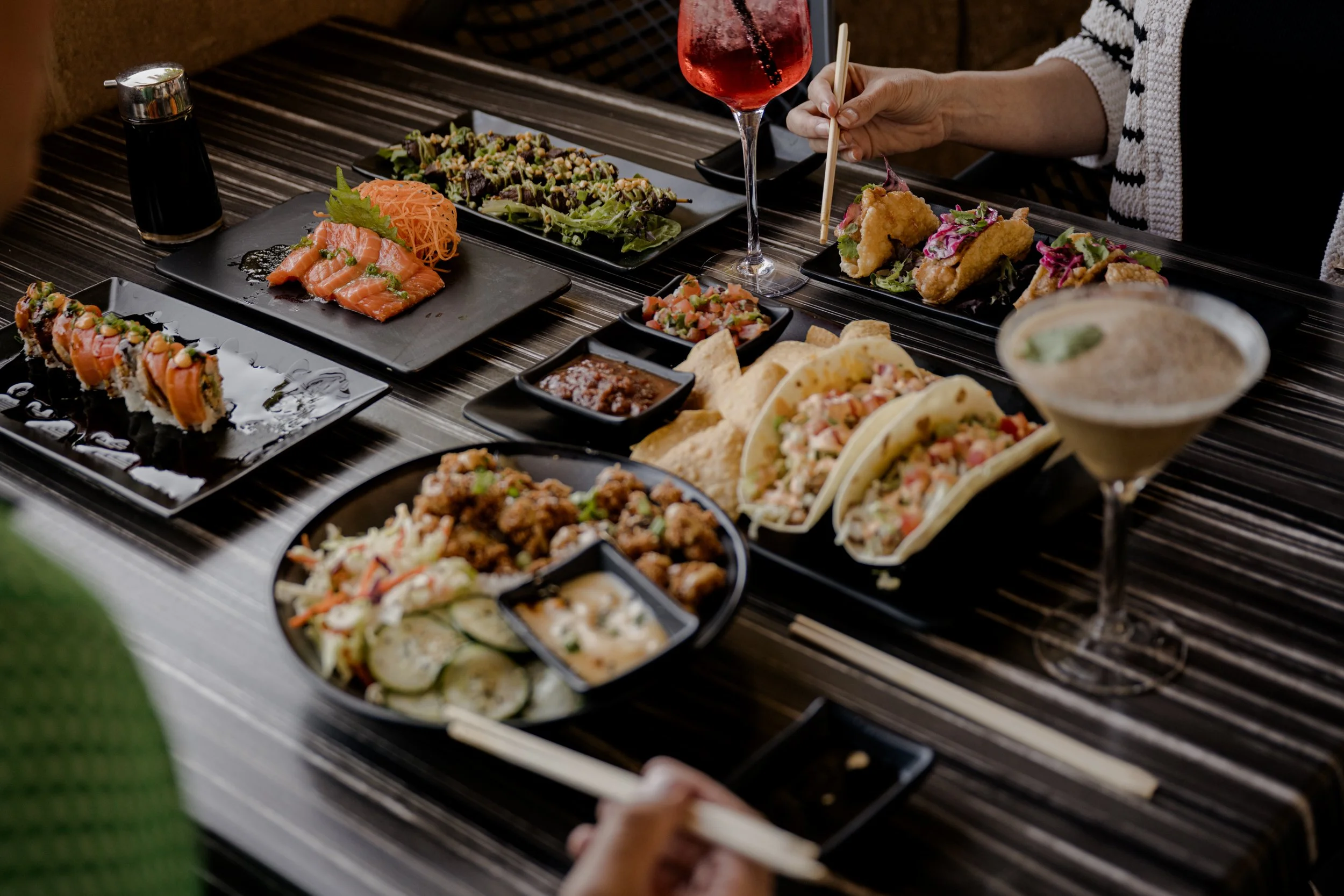 A table set with various Japanese dishes including sashimi, sushi rolls, tacos, fried foods, salads, a cocktail, and other small side dishes, with a person using chopsticks to pick up food.