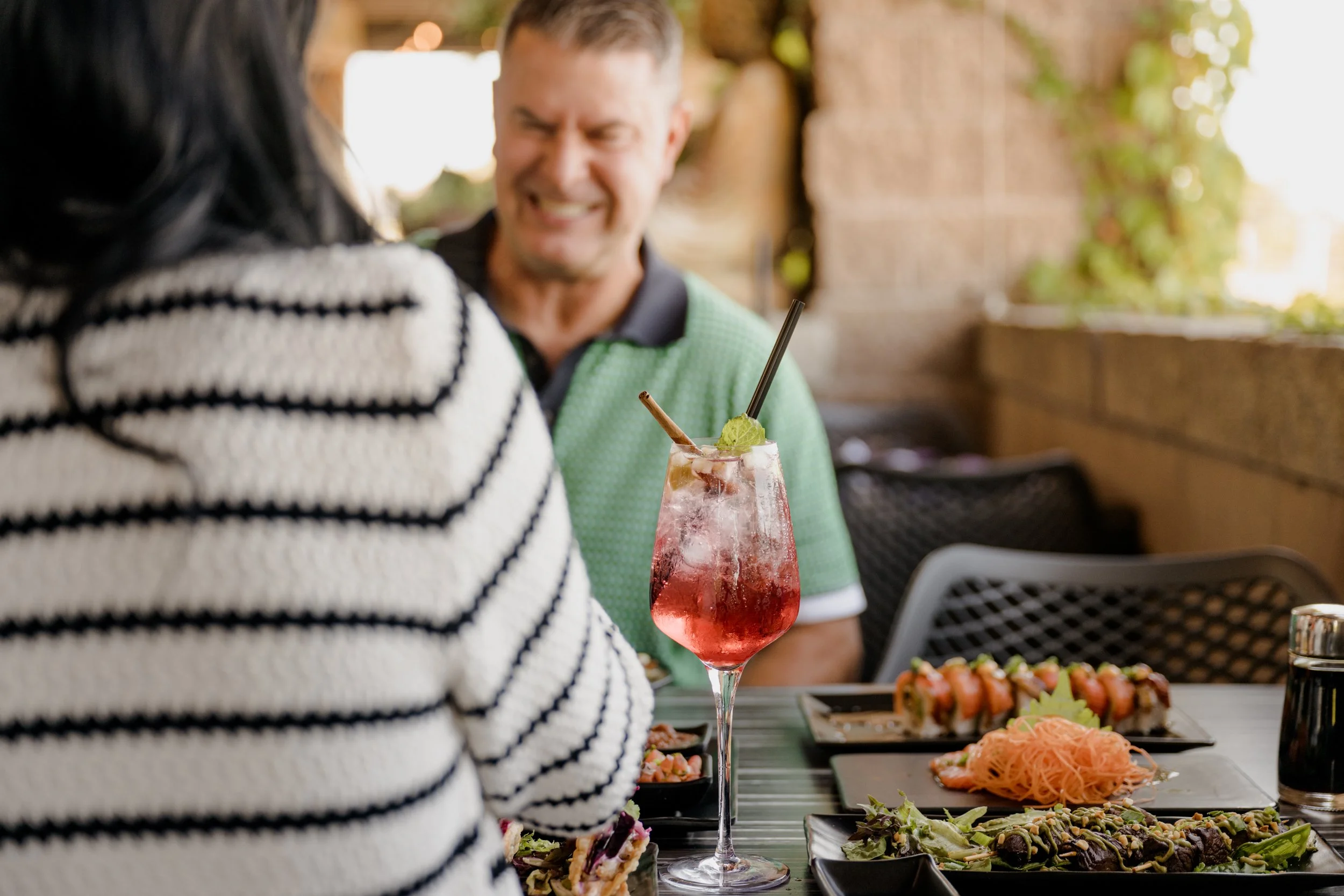 Two people sitting at a table with food and drinks, including a pink cocktail, sushi, and salads, in an outdoor dining area.