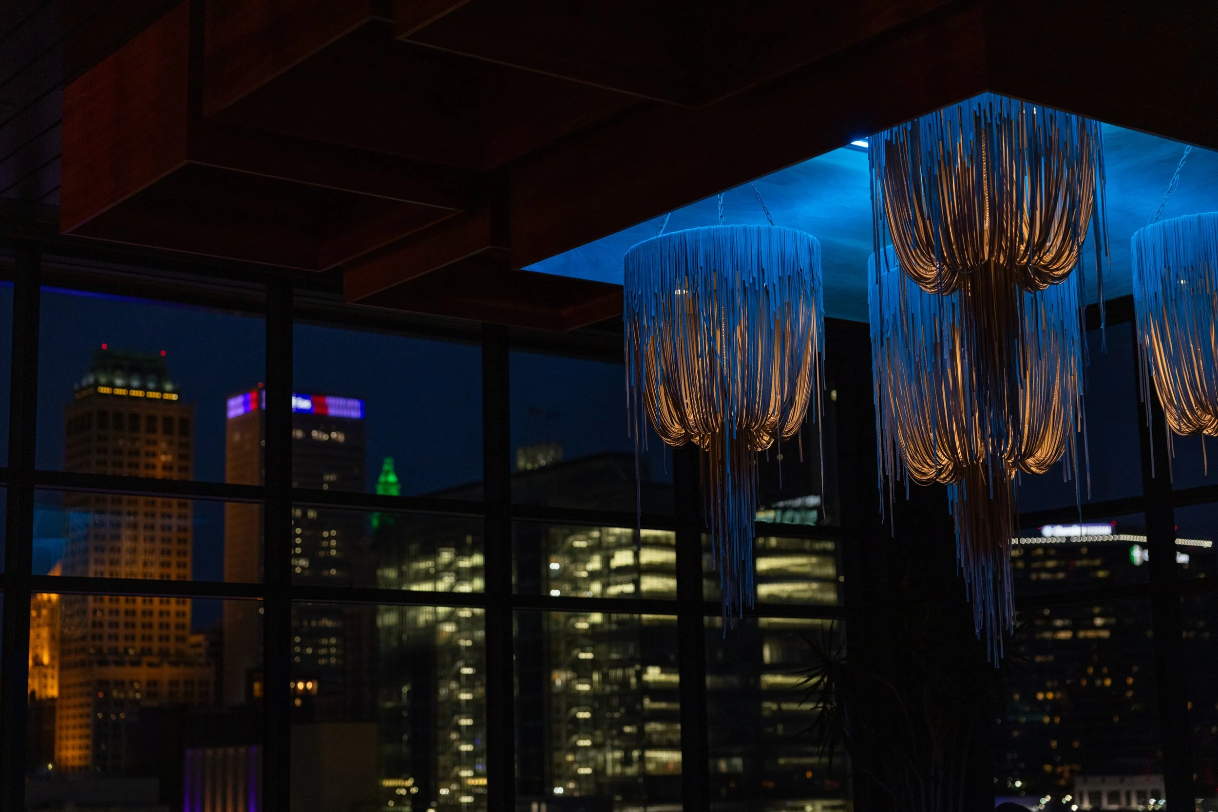 Interior of a modern building at night with city skyline visible through large windows, featuring three hanging light fixtures with fringe design and a cityscape with illuminated skyscrapers in the background.