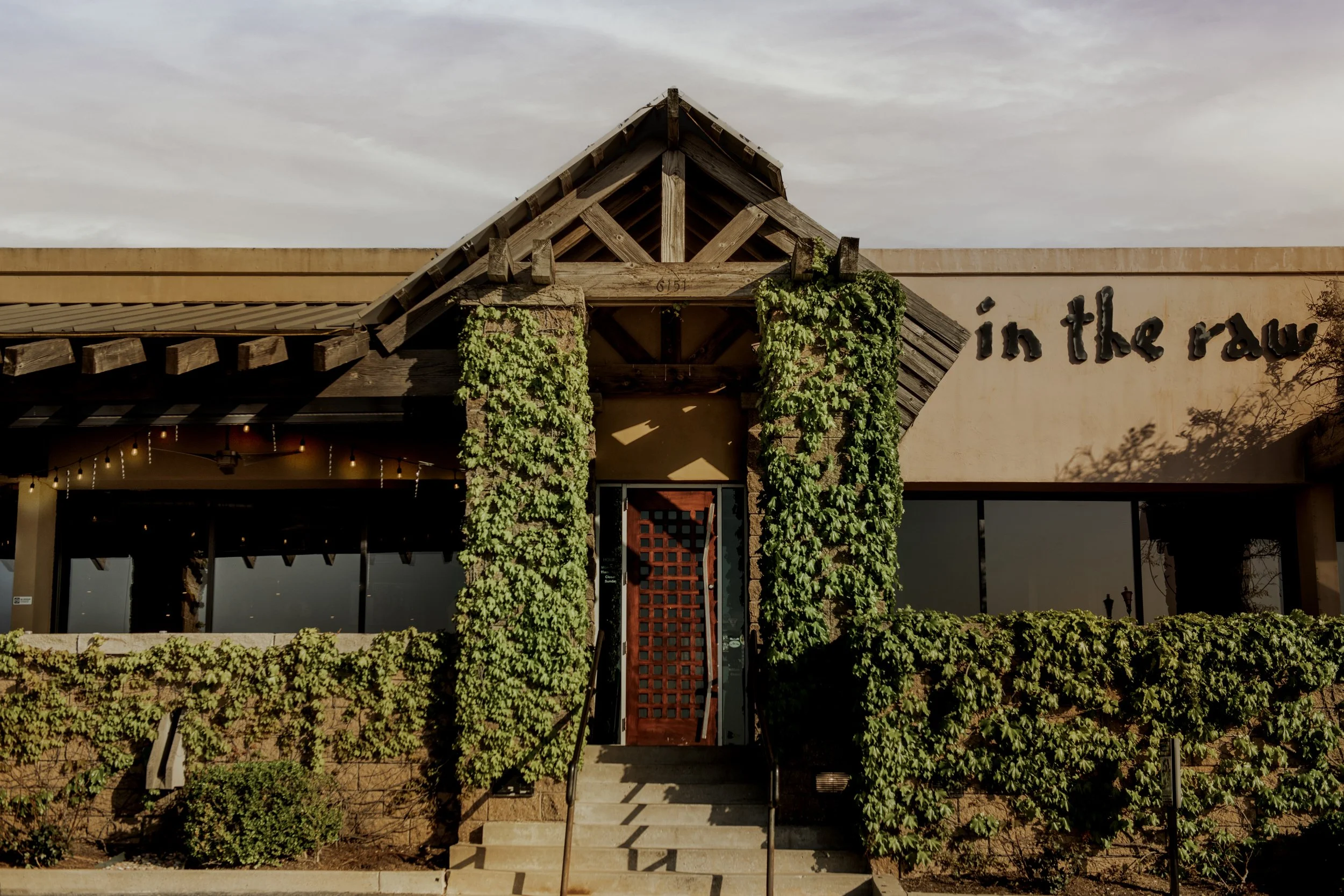 Exterior view of a restaurant with a rustic style, featuring a wooden and stone entrance, greenery on the facade, and a partial sign reading 'in the raw'.