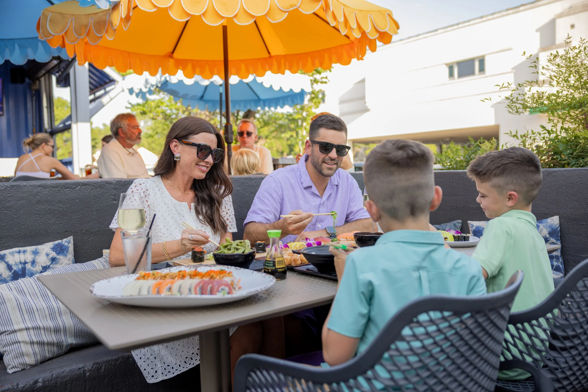 A family enjoying sushi at an outdoor restaurant under colorful umbrellas with other diners in the background.