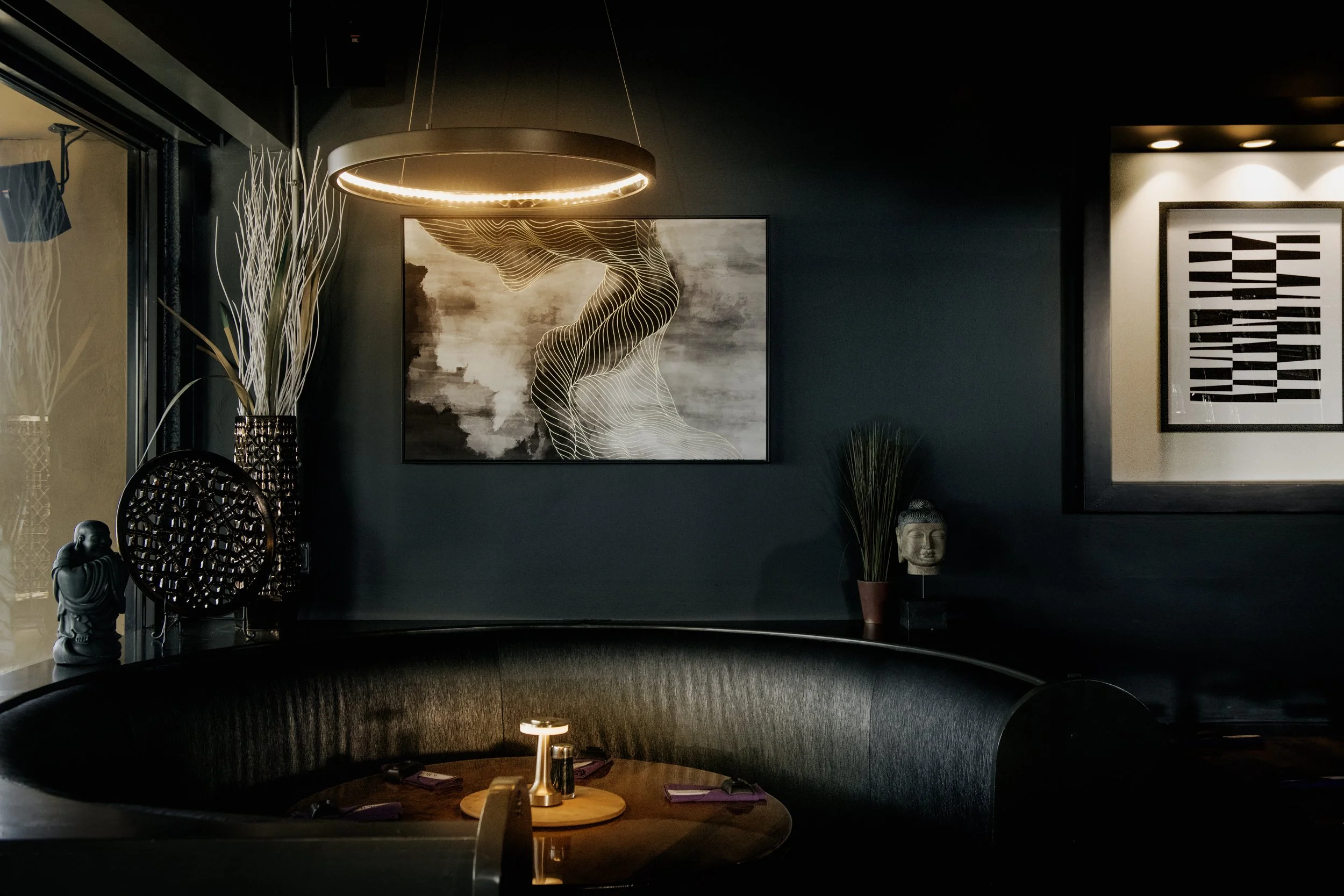 Dark interior of a restaurant or lounge with modern art on the walls, decorative sculptures, and a curved booth with a table set with napkins and small lamps.