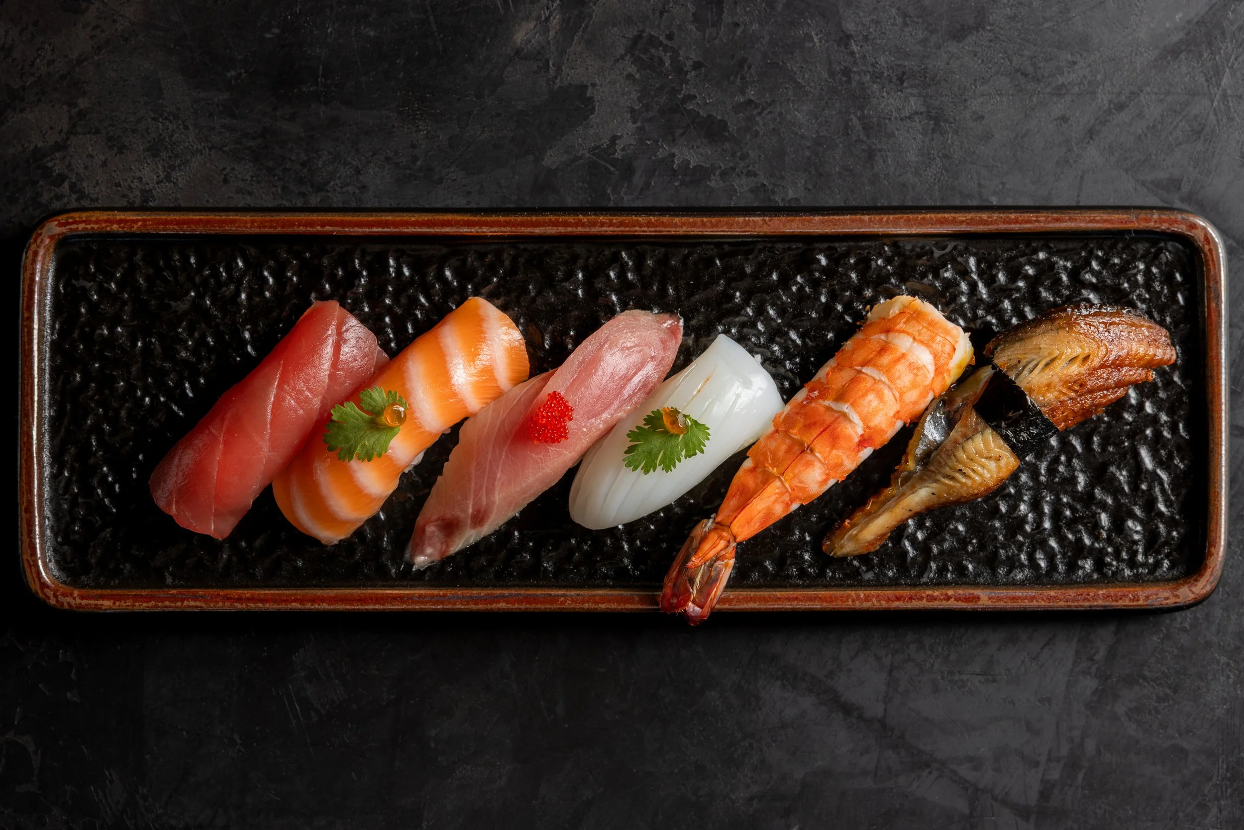 Assorted sashimi sushi on a black textured plate, including tuna, salmon, yellowtail, shrimp, eel, and fish roe garnished with cilantro and wasabi.