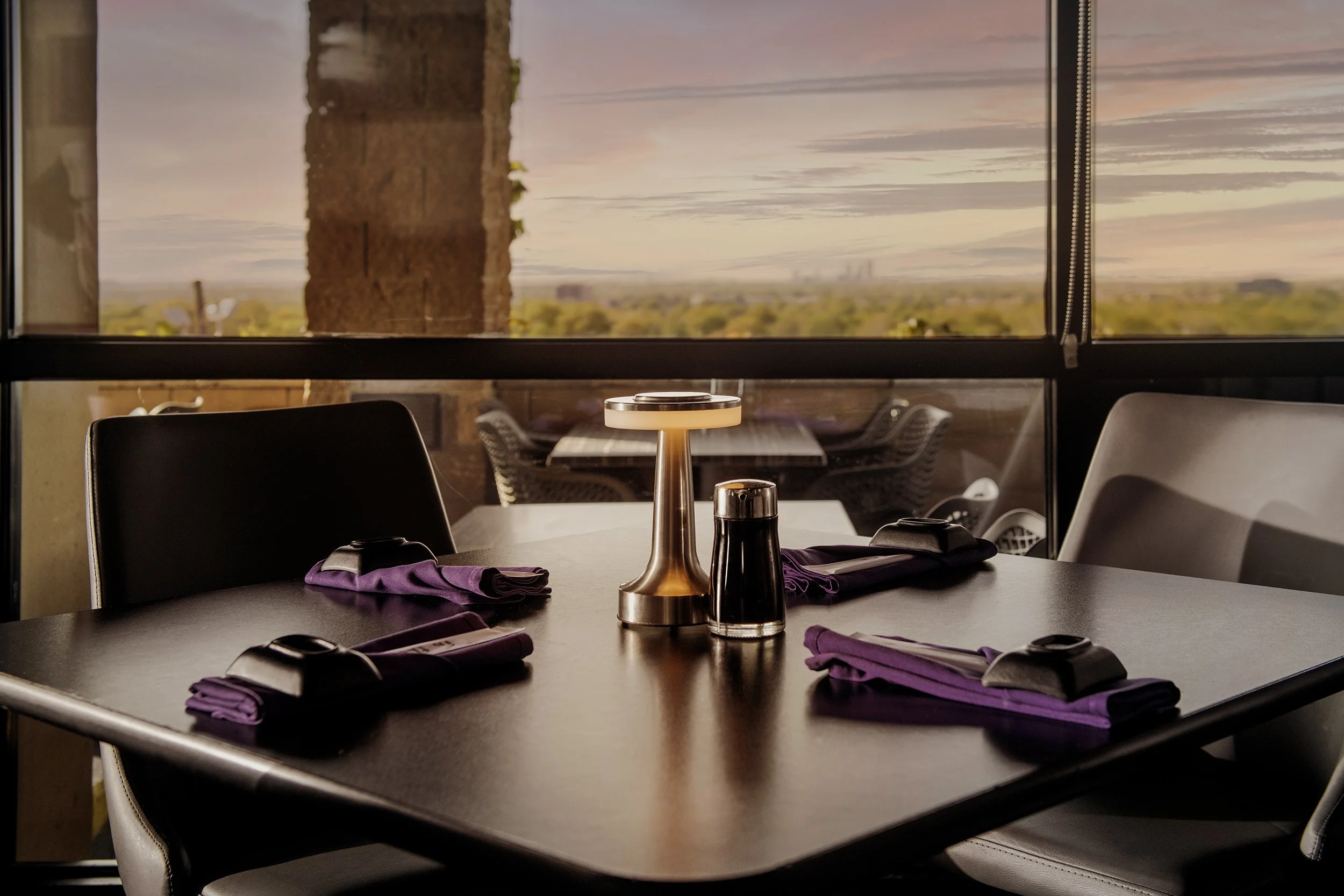 A restaurant table set with purple napkins, black napkin rings, and a modern small lamp, with a view of an outdoor patio and city skyline in the distance at sunset.