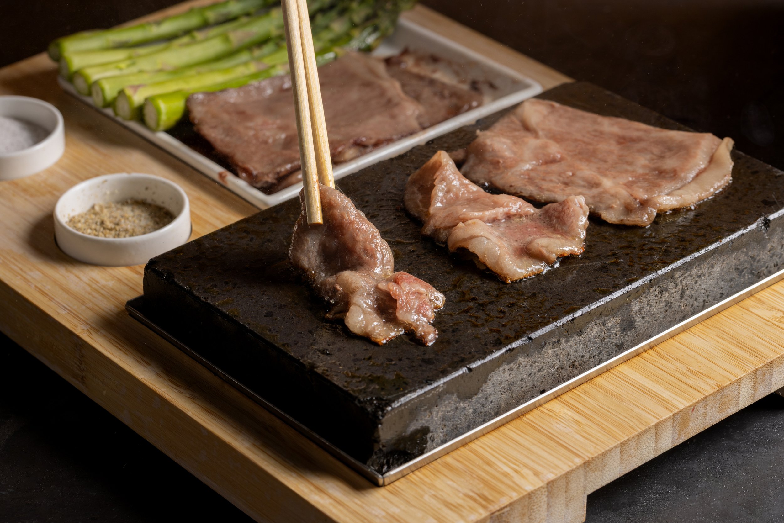Meat and vegetables being cooked on a hot griddle with salt, pepper, and other seasonings in small bowls beside it.
