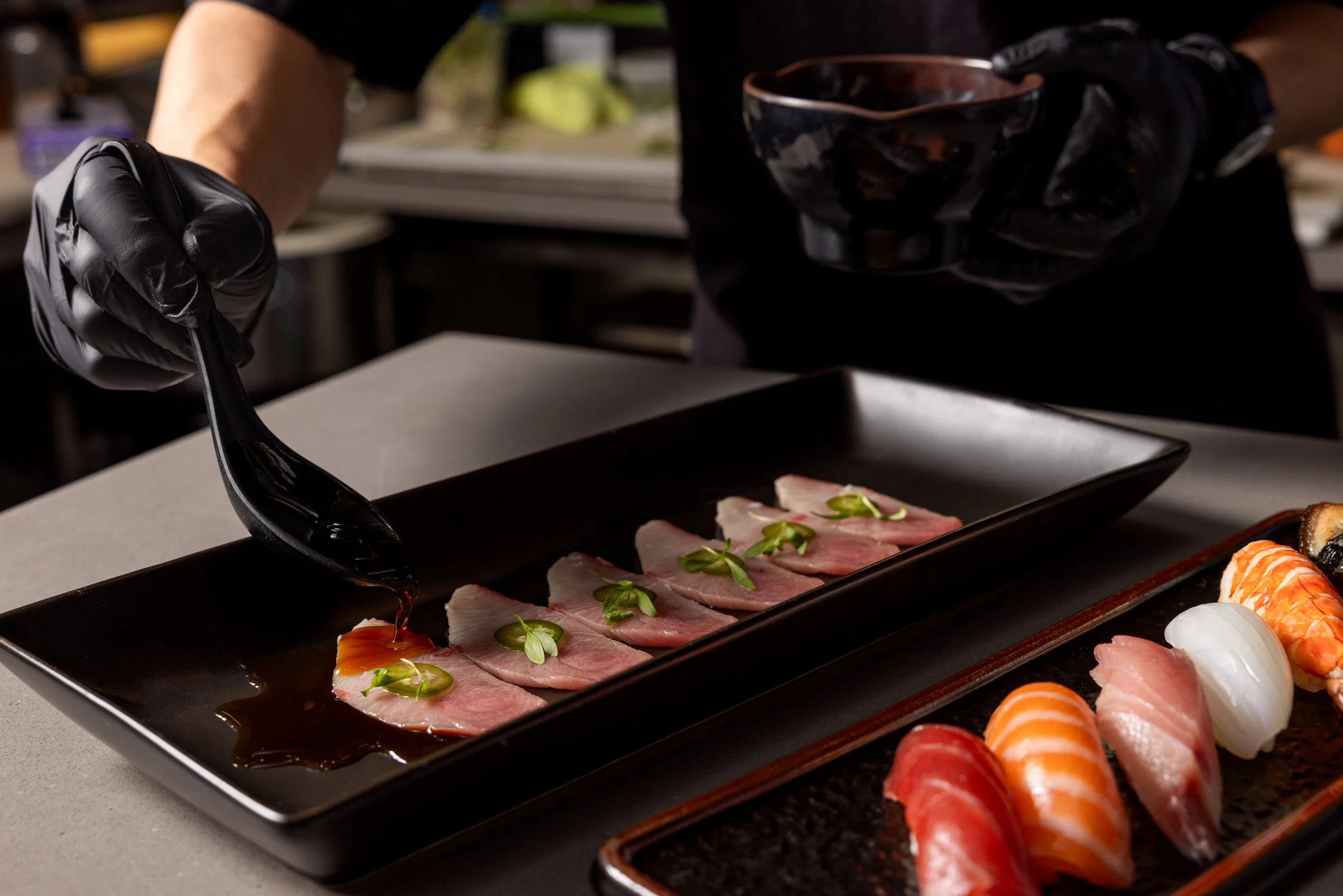 Chef pouring soy sauce over sliced sashimi on a black rectangular plate, with other sushi pieces on a separate serving dish.