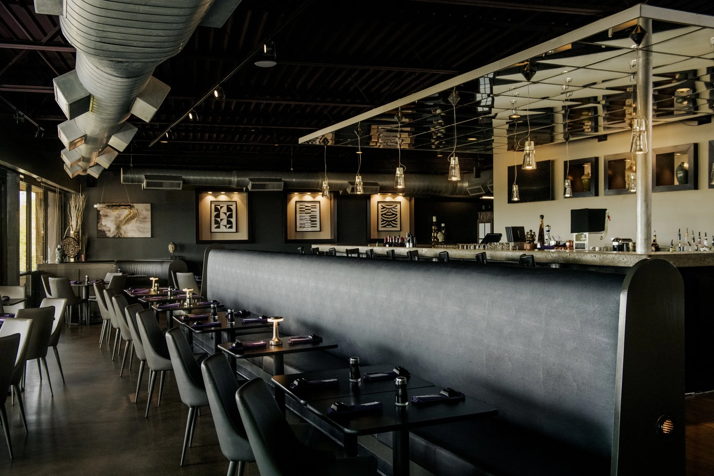 Interior of a modern restaurant or bar with black walls, modular art pieces on the walls, a long bar with a concrete counter, and tables set with black napkins and small candle lights.