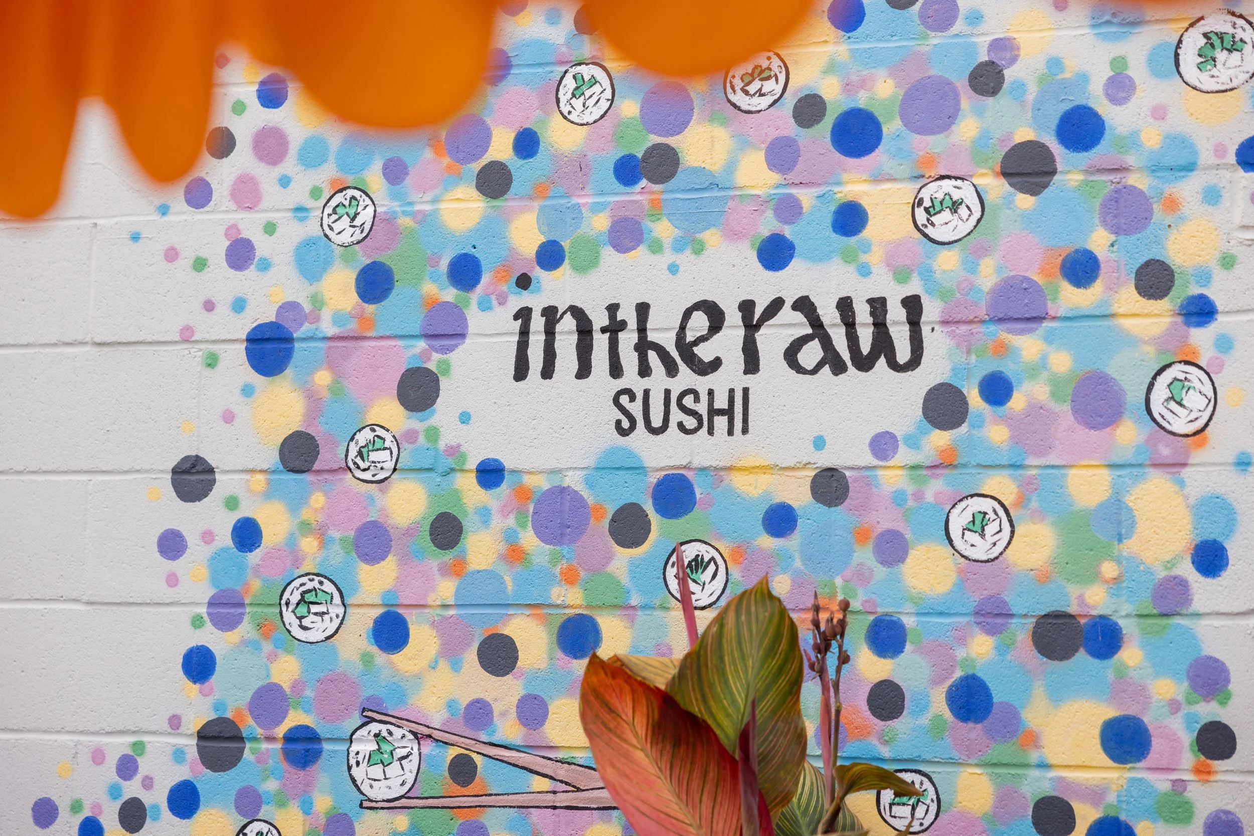 Colorful graffiti on a white brick wall with the words 'inkeraw sushi' in black, surrounded by multicolored dots and fish illustrations, partially obscured by a plant with reddish and green leaves in the foreground.