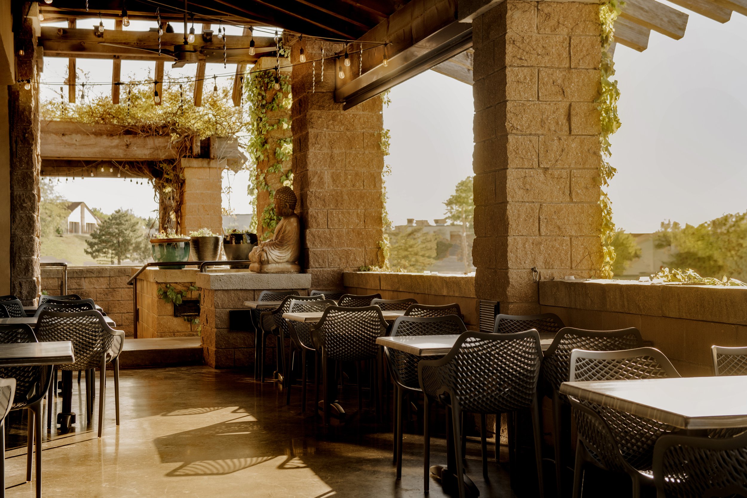 An outdoor dining area with mixed black chairs and tables, a Buddha statue, plants, and string lights on a brick patio during sunset, overlooking a landscape with trees and distant buildings.