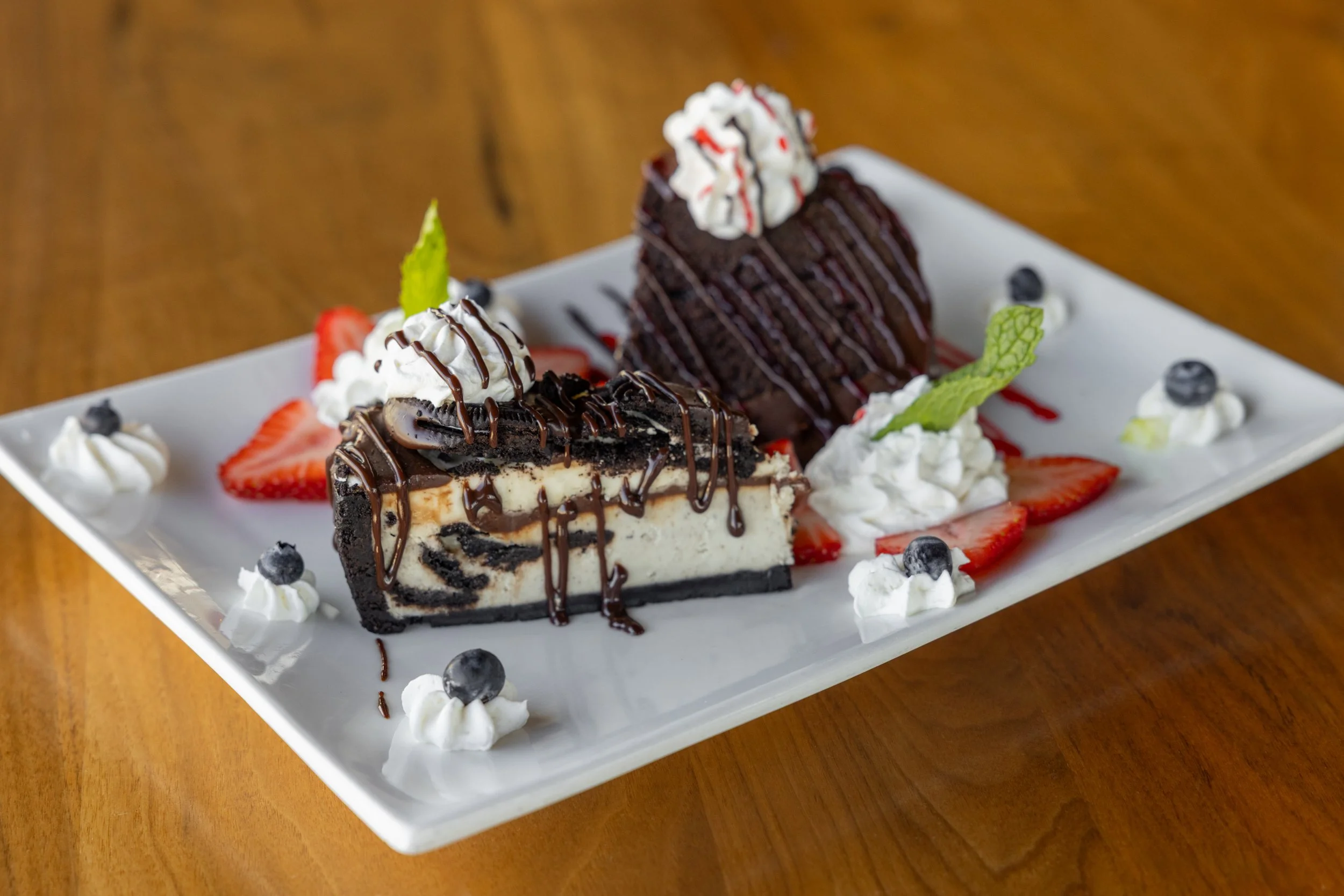 A dessert plate with two slices of cheesecake topped with chocolate and whipped cream, garnished with strawberries, blueberries, and mint leaves, on a white rectangular plate.