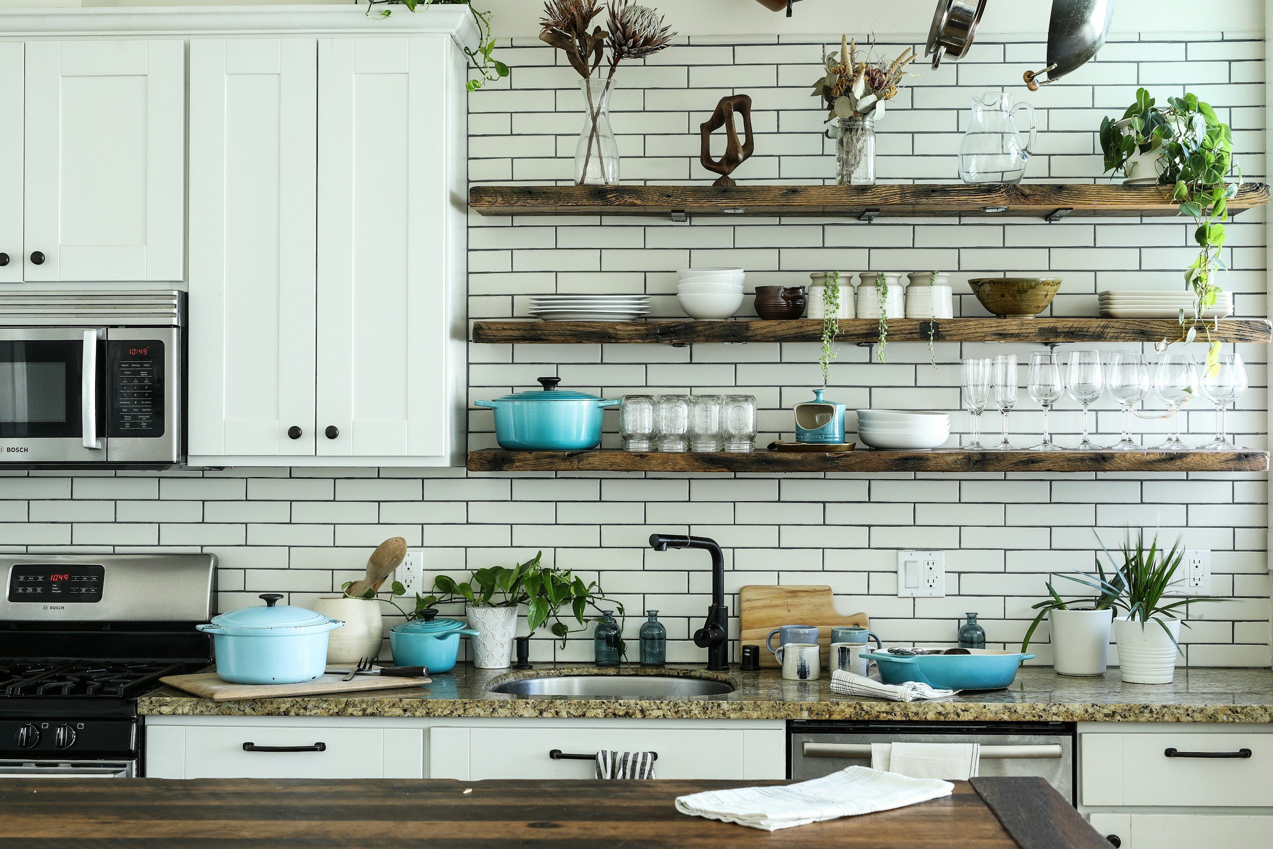 Kitchen with white cabinets, open wooden shelves with dishes and glassware, black faucet, potted plants, and a granite countertop with cooking utensils.