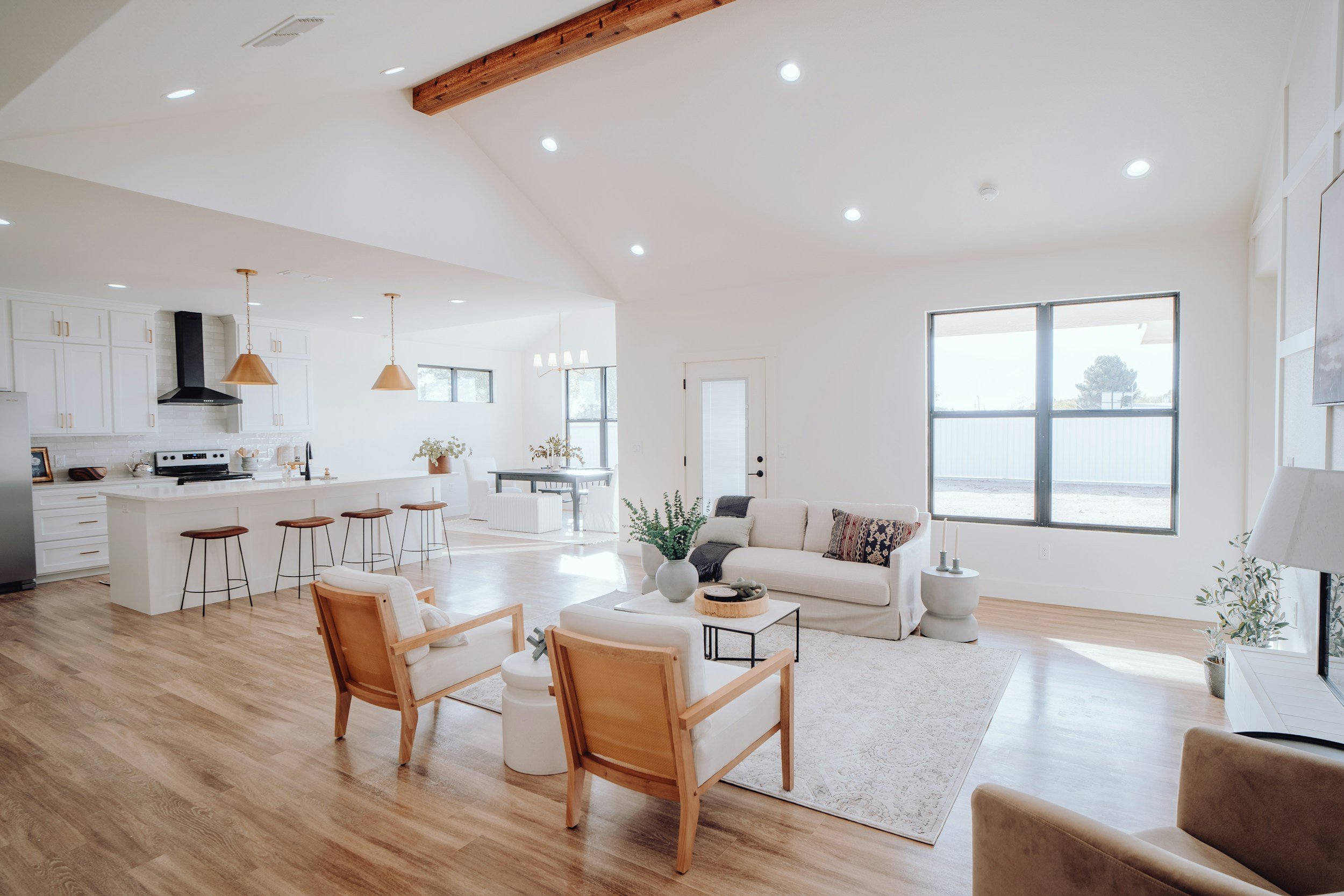 Bright, open-concept living room with white walls, wooden flooring, a white sofa with patterned cushions, two wooden armchairs, a white coffee table, and large windows bringing in natural light.